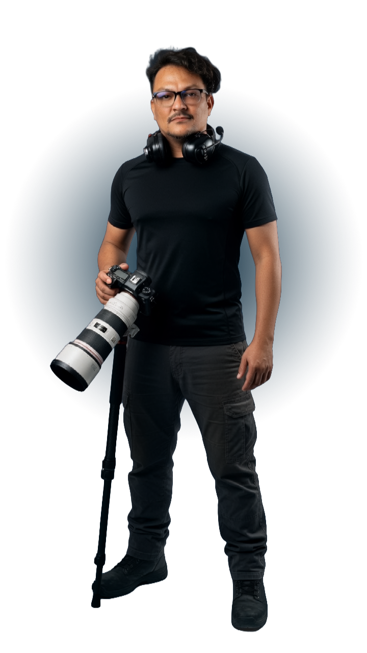 Photographer standing with a large camera lens, wearing a black t-shirt, cargo pants, and headphones around neck, against a dark background.