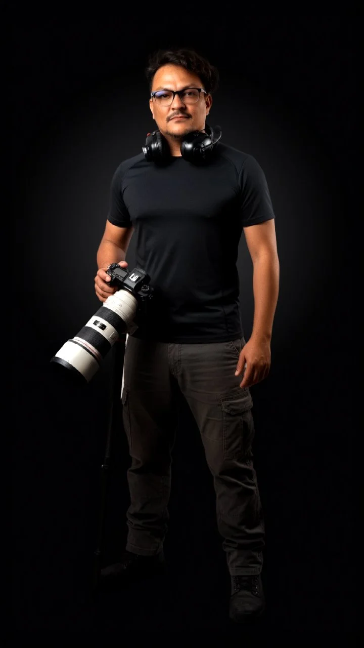 Man wearing glasses with headphones around neck, holding a camera with a large telephoto lens, standing against a black background