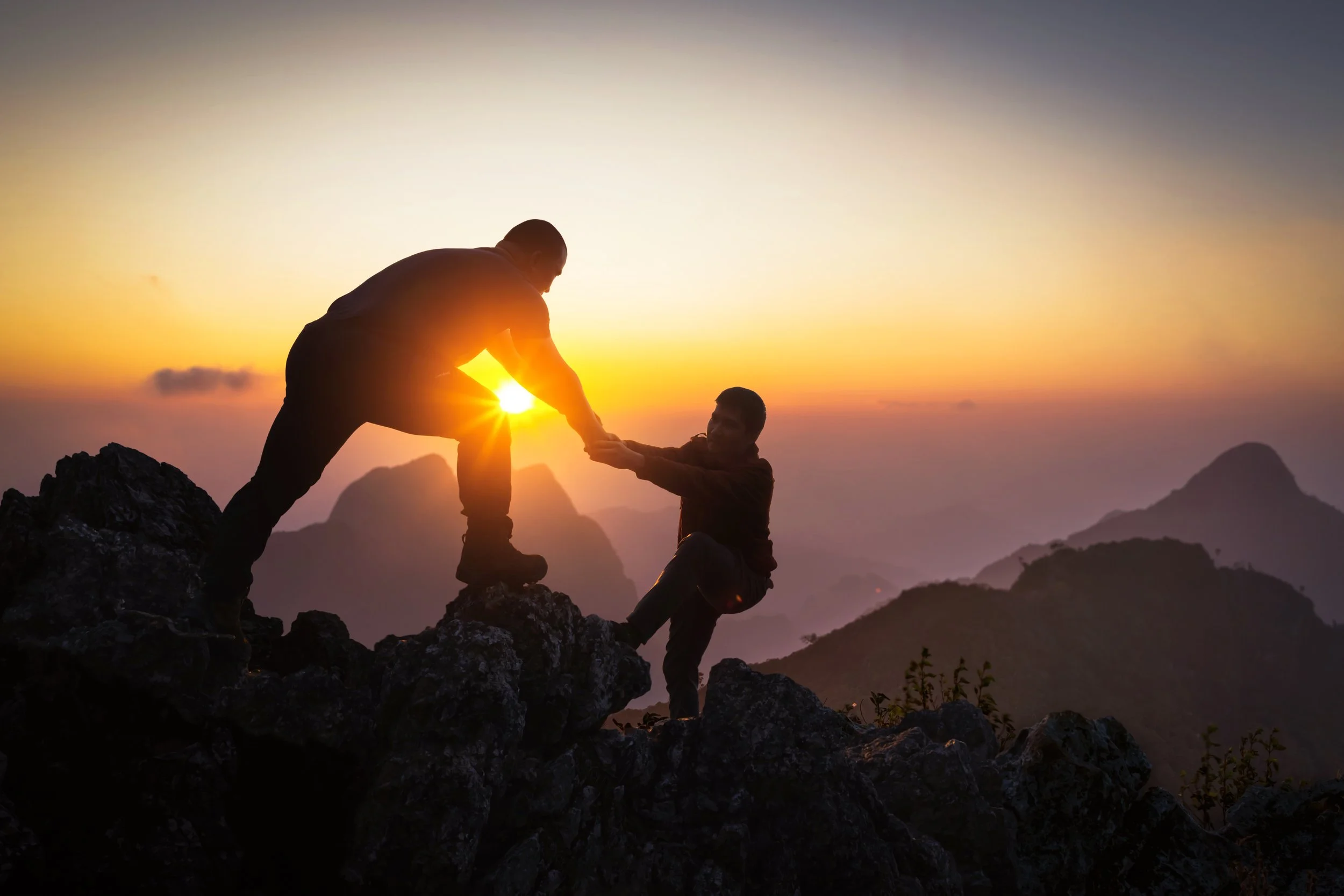 One person reaching back to help another climb a mountain at sunrise