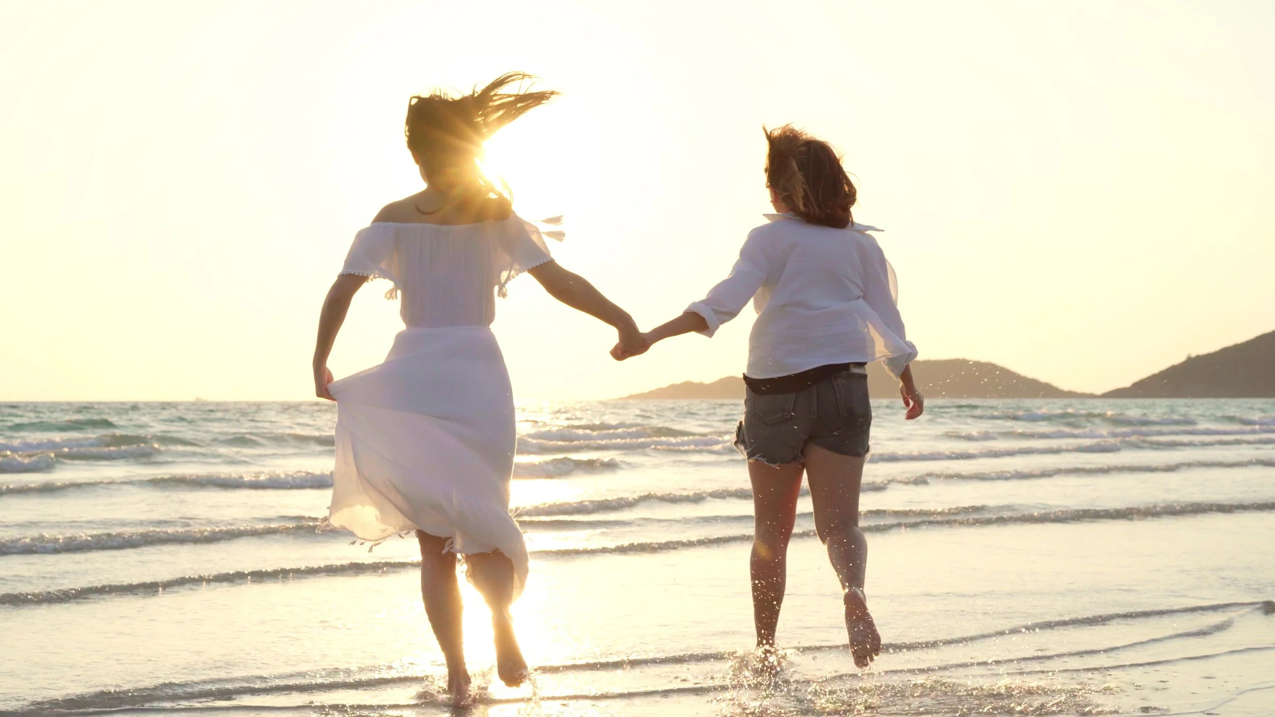 Two women running together along the beach at sunset, holding hands