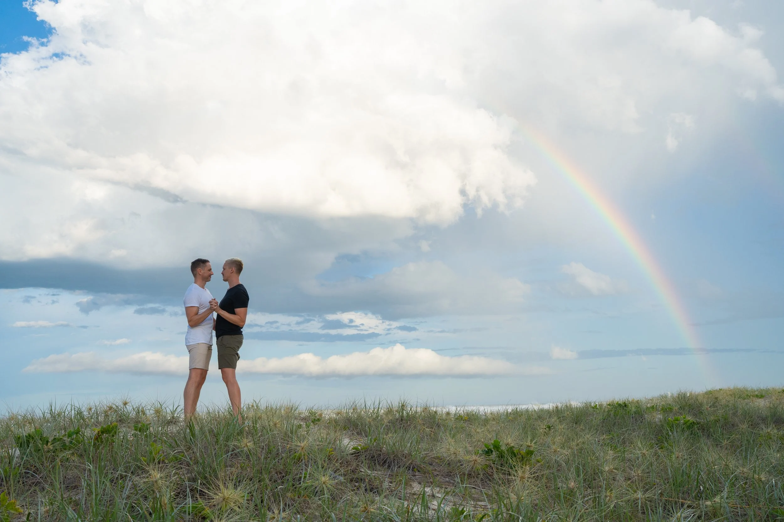 Same-sex couple embracing on the beach beneath a rainbow