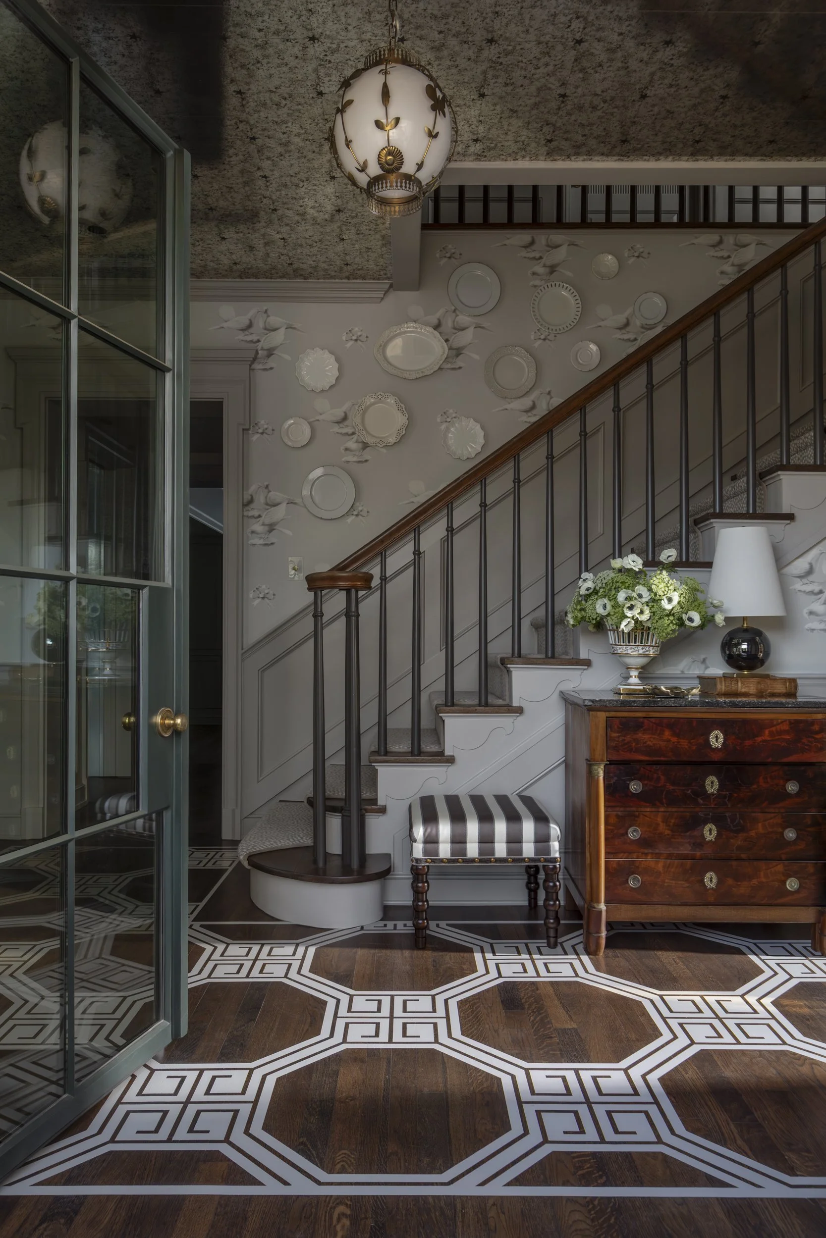 Interior of a house featuring a staircase with black spindles and wooden handrail, decorated white wall plates and sculptures, a vintage wooden dresser with a flower vase, a striped upholstered stool, and a patterned tile floor.