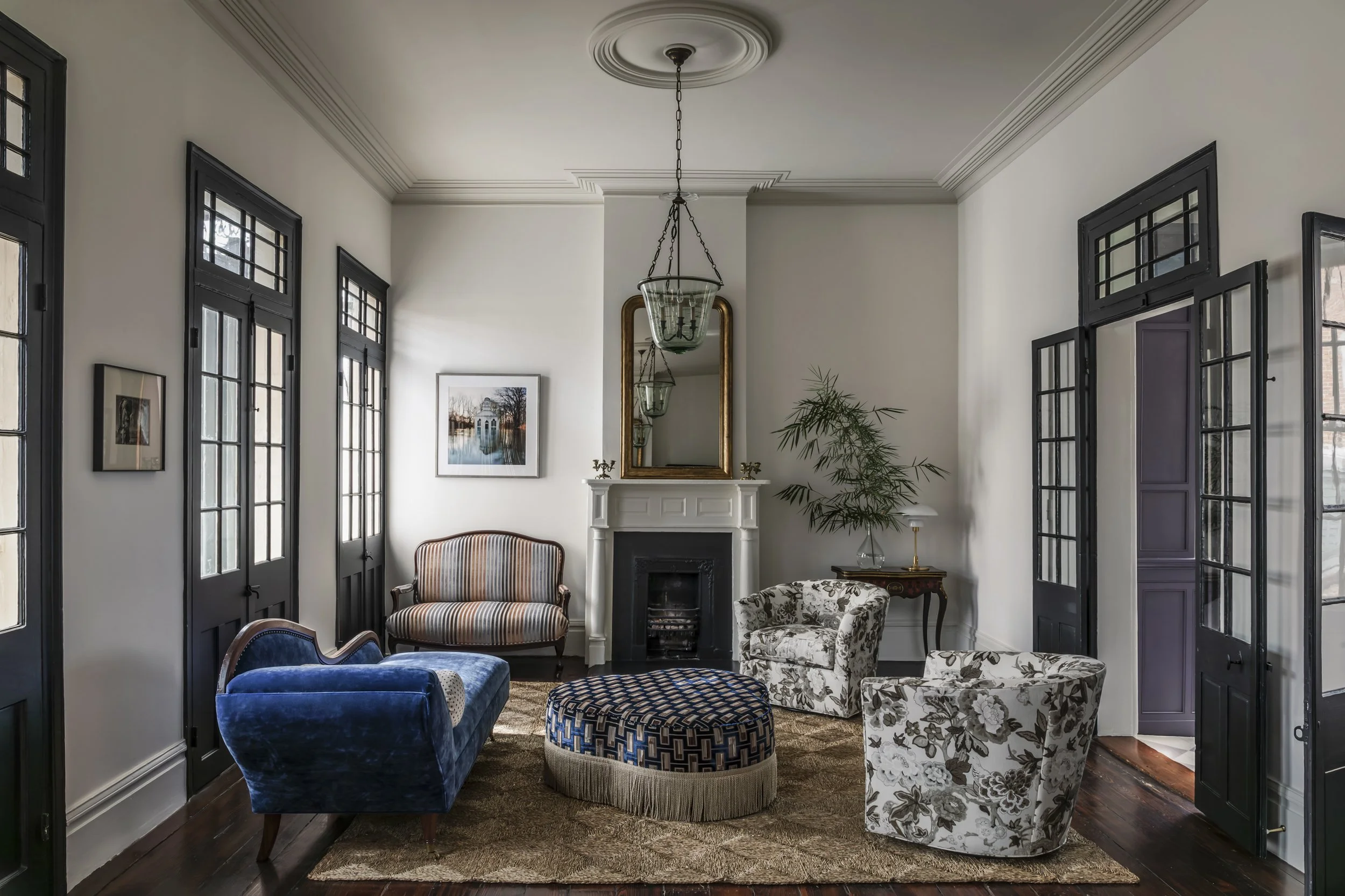 French Quarter parlor with high ceilings, white walls, and black window frames. Furniture includes a blue velvet sofa, patterned armchairs, and a round ottoman. An ornate antique mirror hangs above a fireplace, and framed artwork decorates the walls.