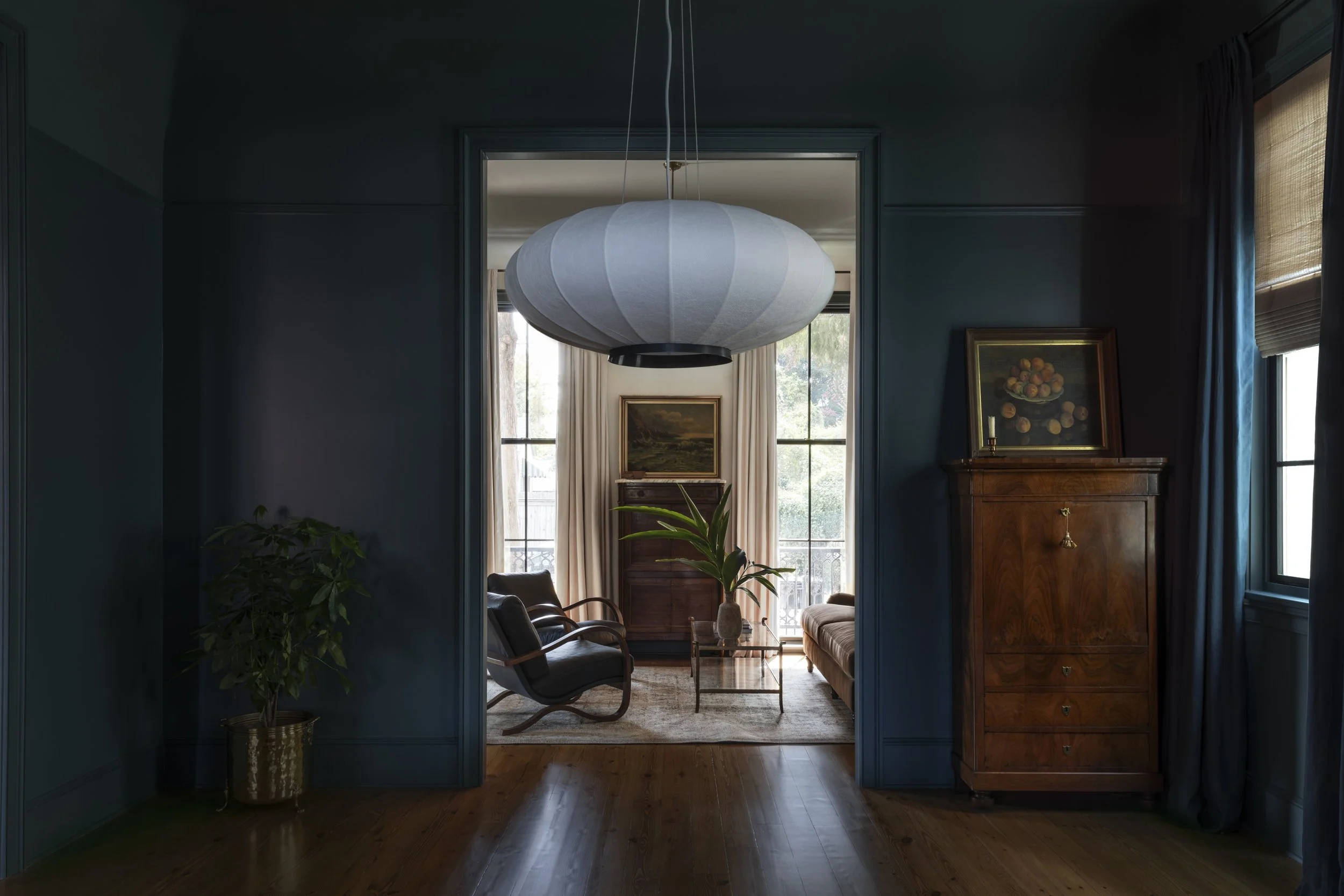 Interior view of a living room featuring blue walls, a large white round paper lantern hanging from the ceiling, a wooden cabinet with a framed painting on top, and windows with curtains allowing natural light to fill the space.