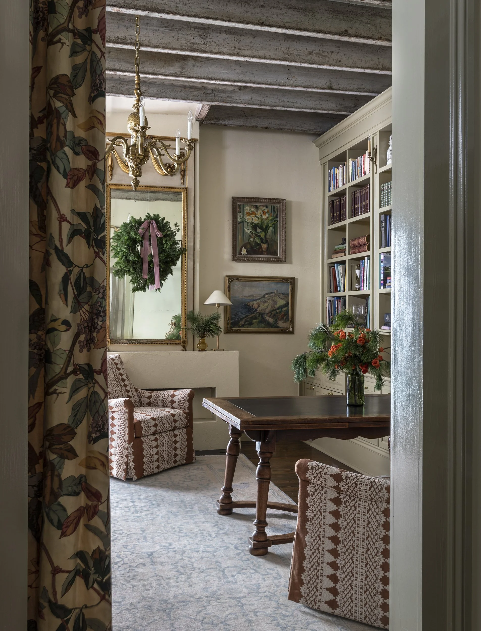 A cozy living room with a floral curtain partially pulled back, revealing a wooden dining table with a vase of flowers, checkered armchairs, a white bookshelf filled with books, framed artwork on the wall, a brass chandelier, and a wreath hanging on 