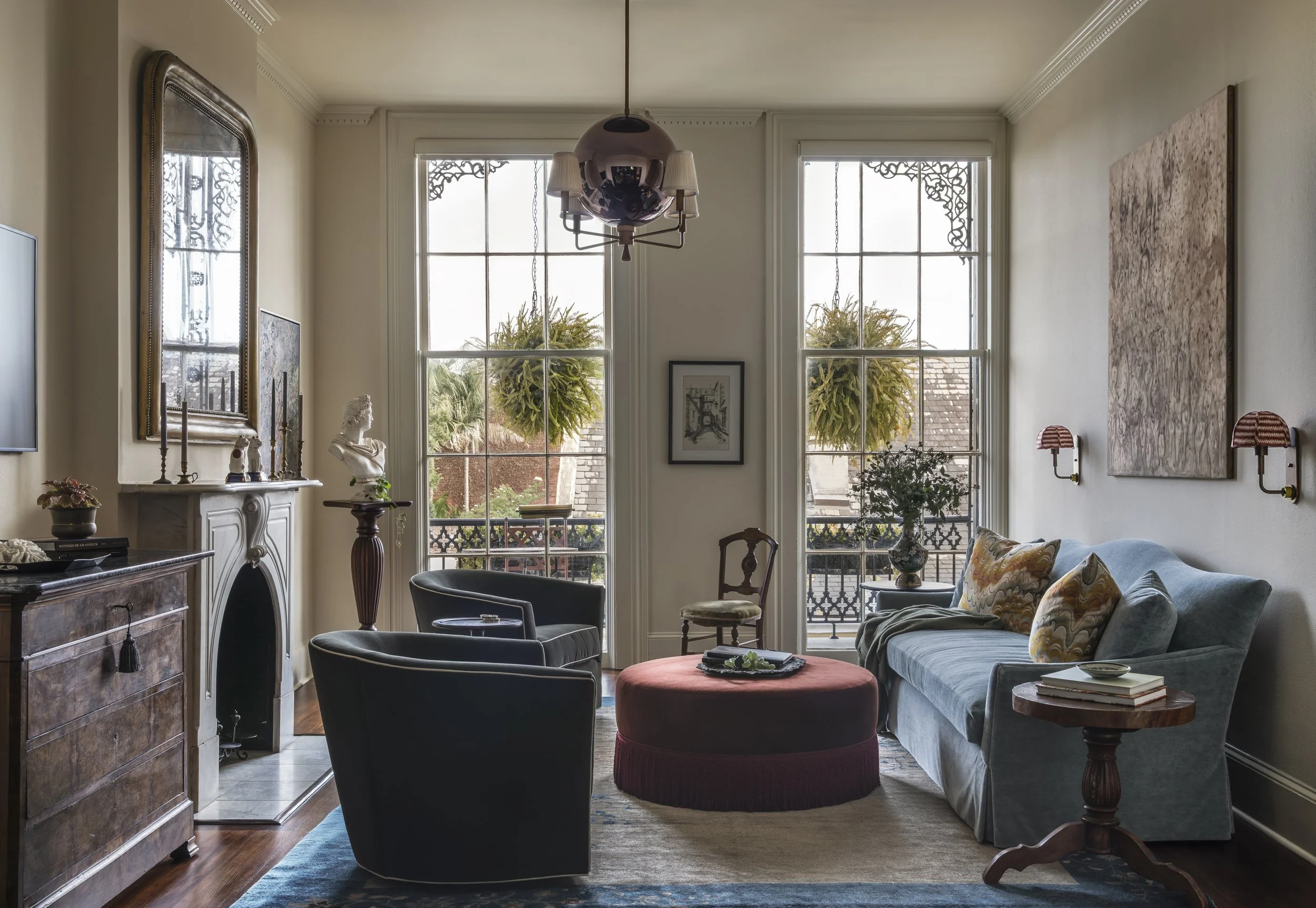 Living room with large windows, eclectic furniture including a blue sofa with patterned pillows, black armchairs, wooden side tables, and various art pieces and decorations. There are plants hanging outside the windows, and a chandelier hanging from 