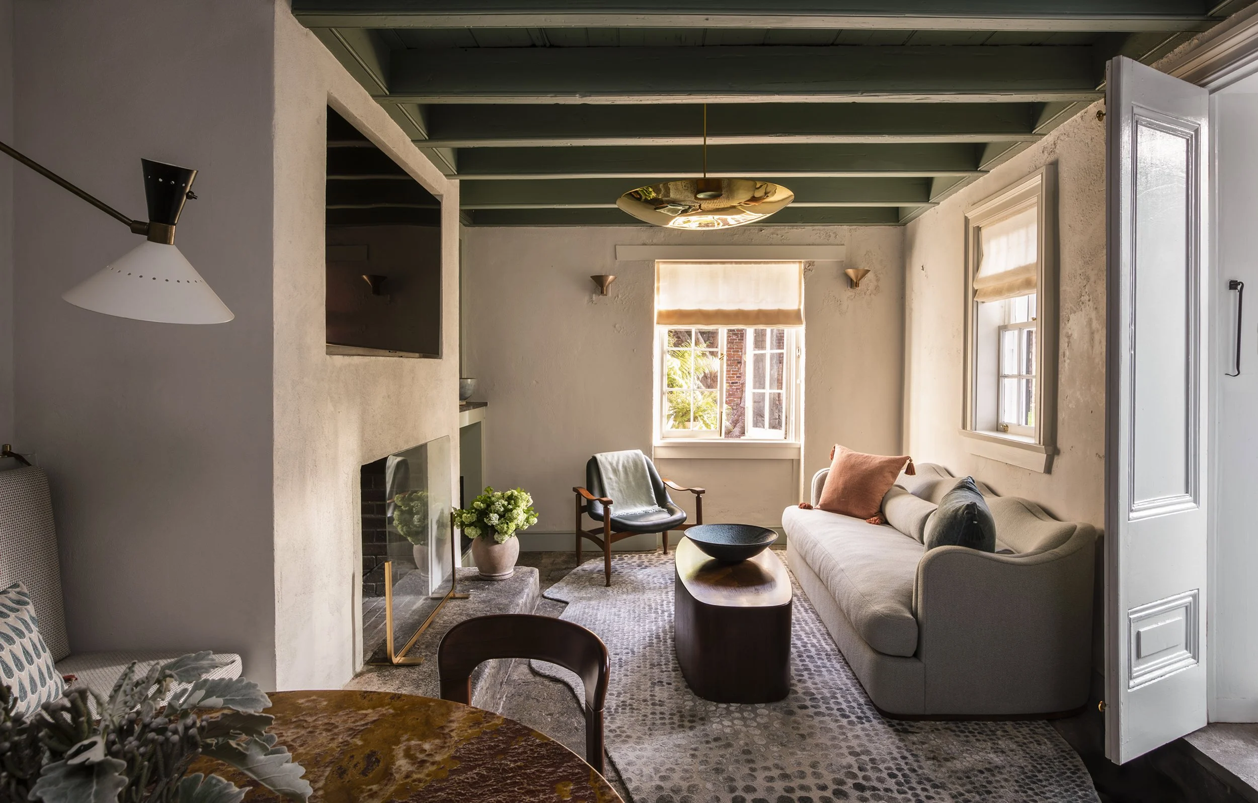 A cozy living room with a beige sofa, a black armchair, and a wooden coffee table. There are multiple windows with natural light, a potted plant with white flowers, and a patterned rug on the floor. The ceiling has green beams and a gold ceiling ligh
