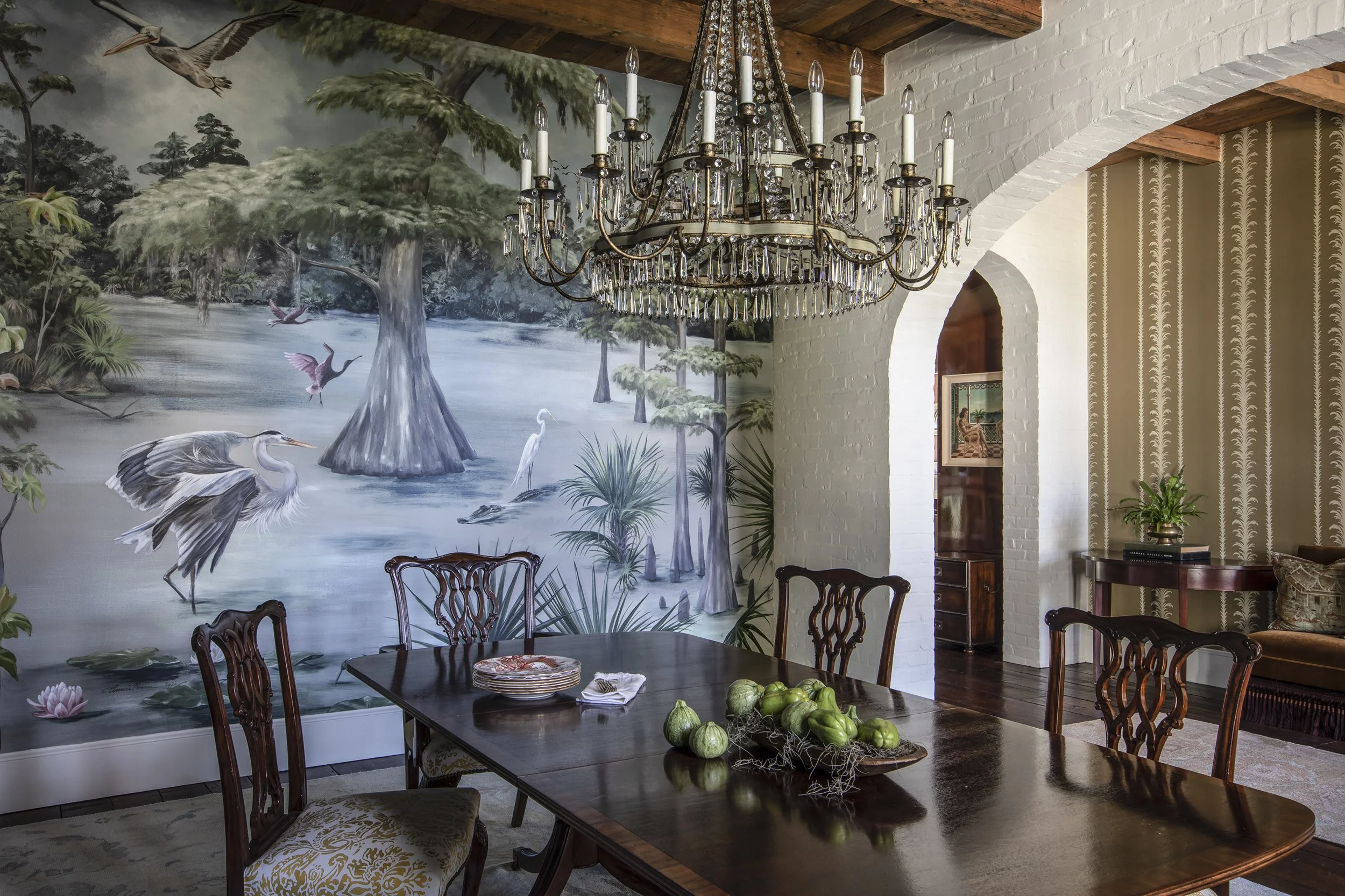 Interior of a dining room with a large mural of a swamp scene featuring herons and cypress trees on the wall. A grand chandelier with crystal accents hangs from a wooden beamed ceiling. The room has white painted brick walls with an arched doorway, p