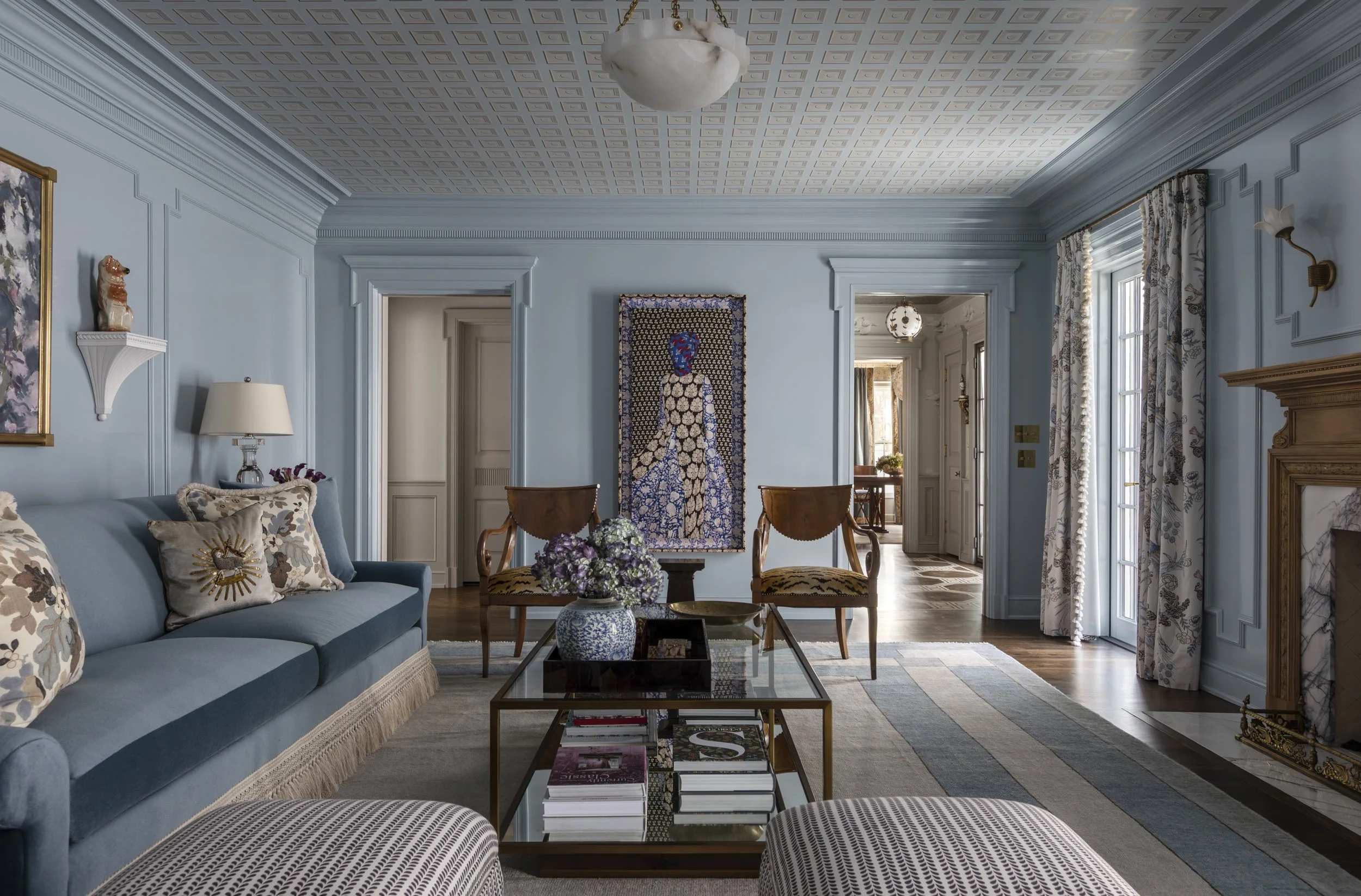 A living room with light blue walls, a blue sofa with patterned pillows, a glass coffee table with magazines, and natural light from tall windows with patterned curtains. There's artwork on the walls and a floral arrangement on the coffee table.