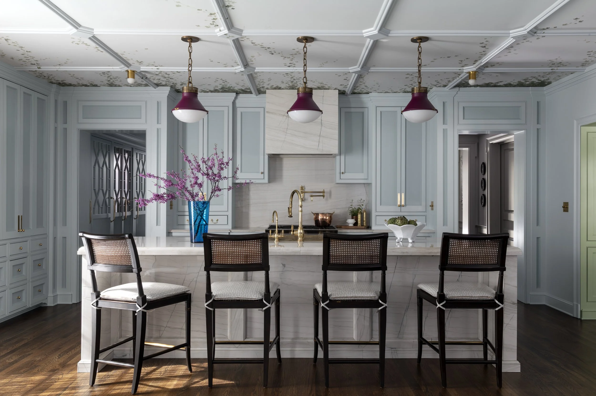 A modern kitchen with a large white island, black chairs, light blue cabinets, marble backsplash, and a ceiling with decorative molding and three pendant lights with burgundy shades. A blue vase with pink flowers and a bowl of green vegetables are on