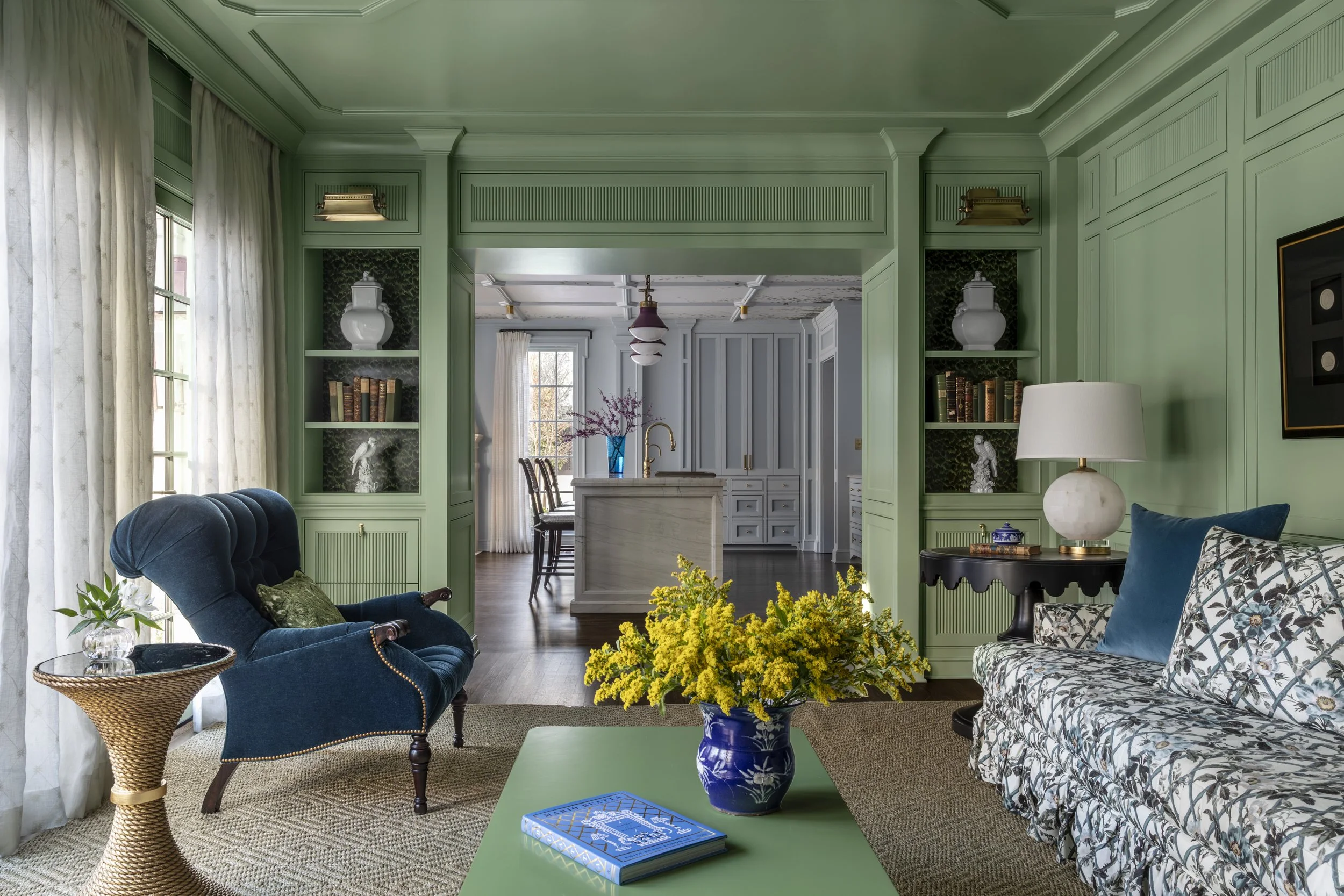 Interior of a living room with a green wall, dark blue armchair, floral sofa, green coffee table with a blue book, yellow flowers in a blue vase, and window with white curtains. In the background, a kitchen with gray cabinetry and a dining area with 