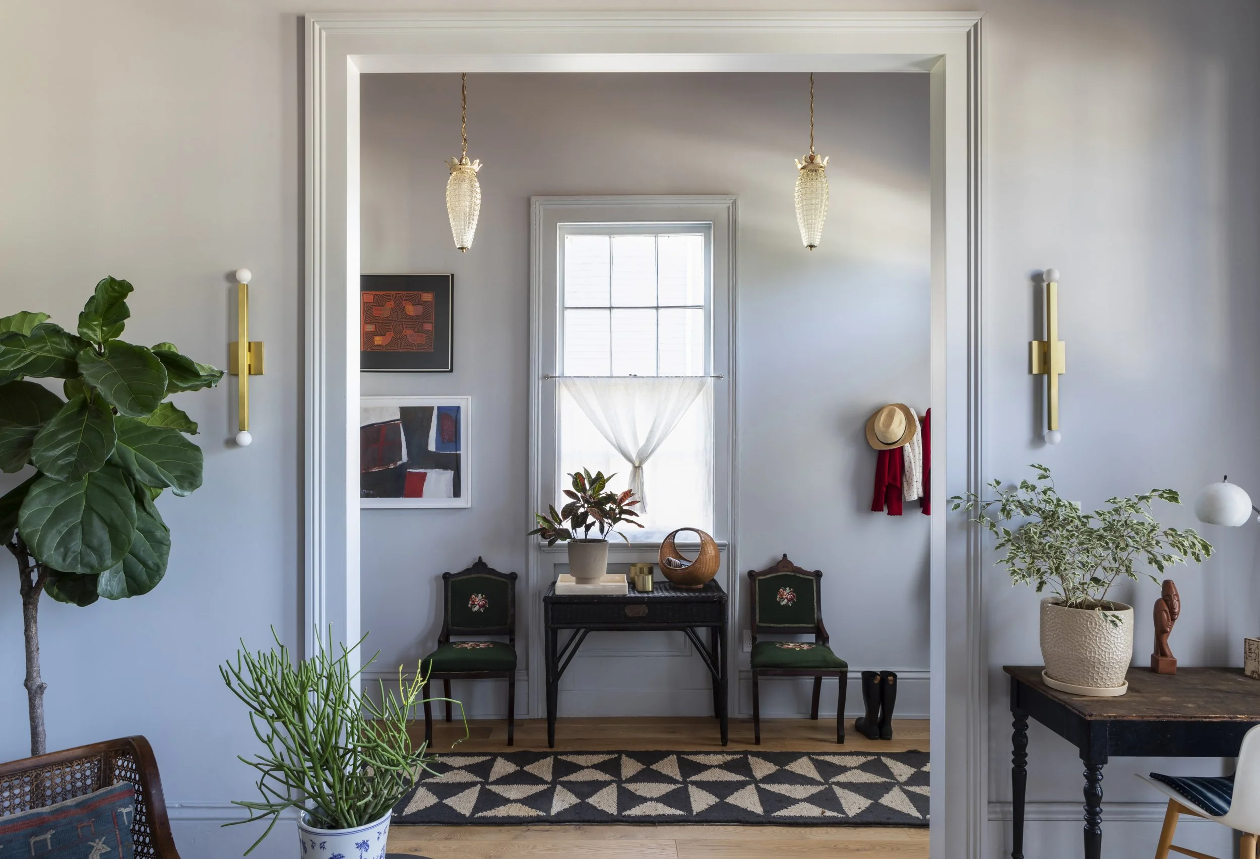 A living room or dining area with an open doorway leading to a bright room with a window. The space features artwork on the walls, two vintage chairs with green upholstery, an ornate console table, and various decorative items including potted plants
