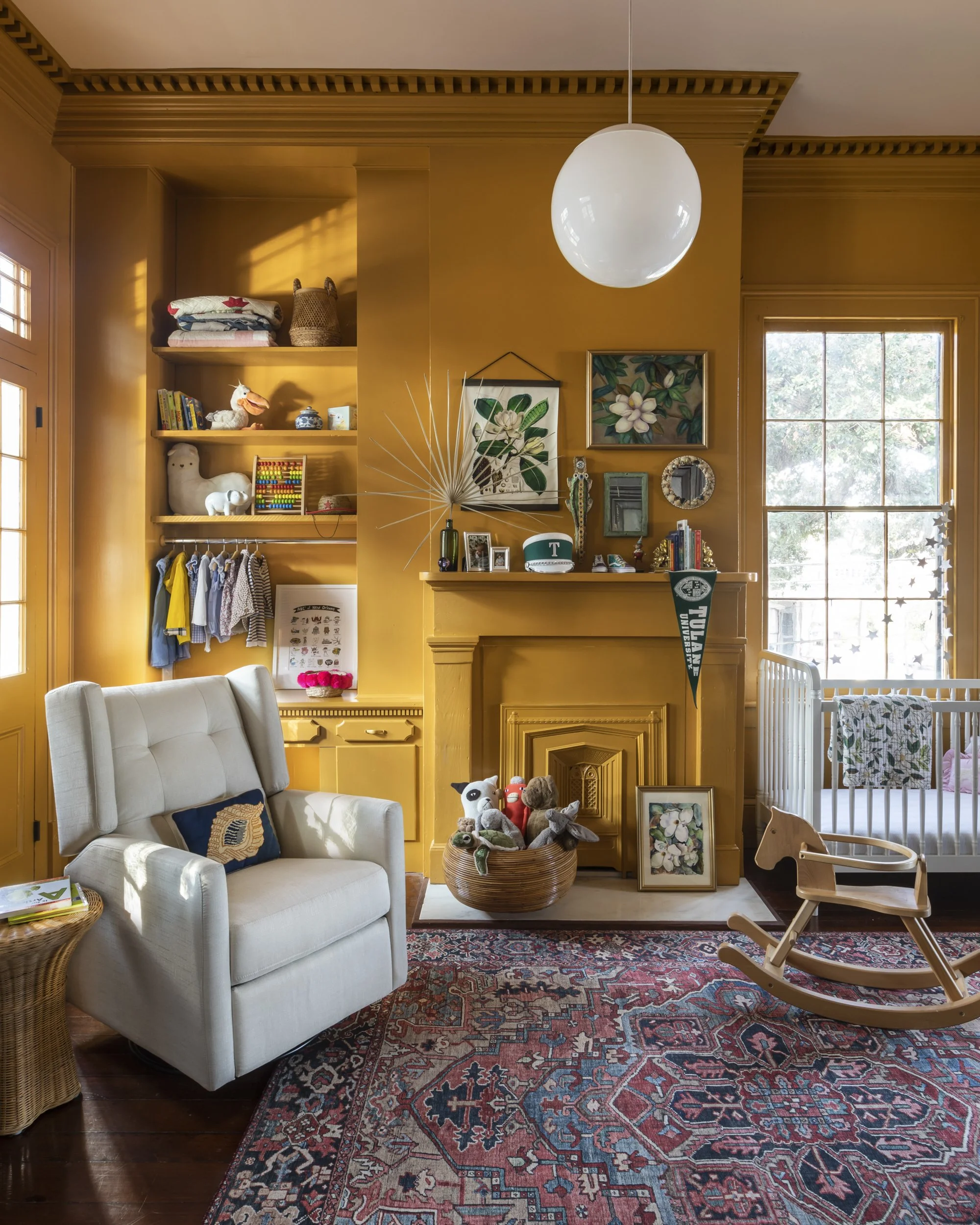 A cozy living room with mustard yellow walls and decorative wooden crown molding. There is a white armchair with a patterned pillow, a colorful Persian rug, and a wicker basket filled with children's stuffed toys. On the wall, there are framed botani