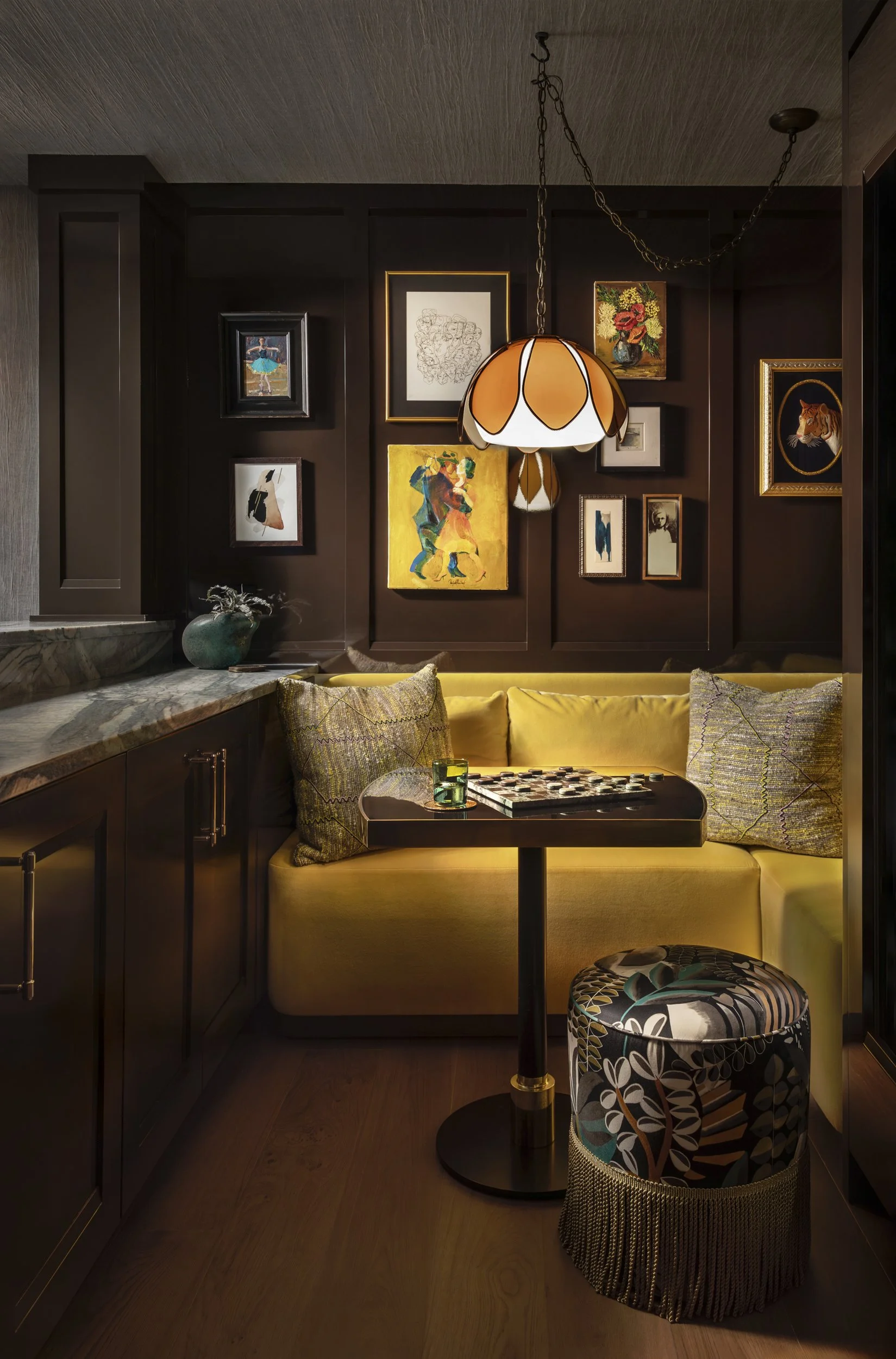 A cozy corner of a room with dark wall adorned with framed artwork, including a horse, a tiger, a dancer, a vase with flowers, and abstract pieces, illuminated by a hanging lamp with a petal design, next to a yellow sofa with patterned cushions and a