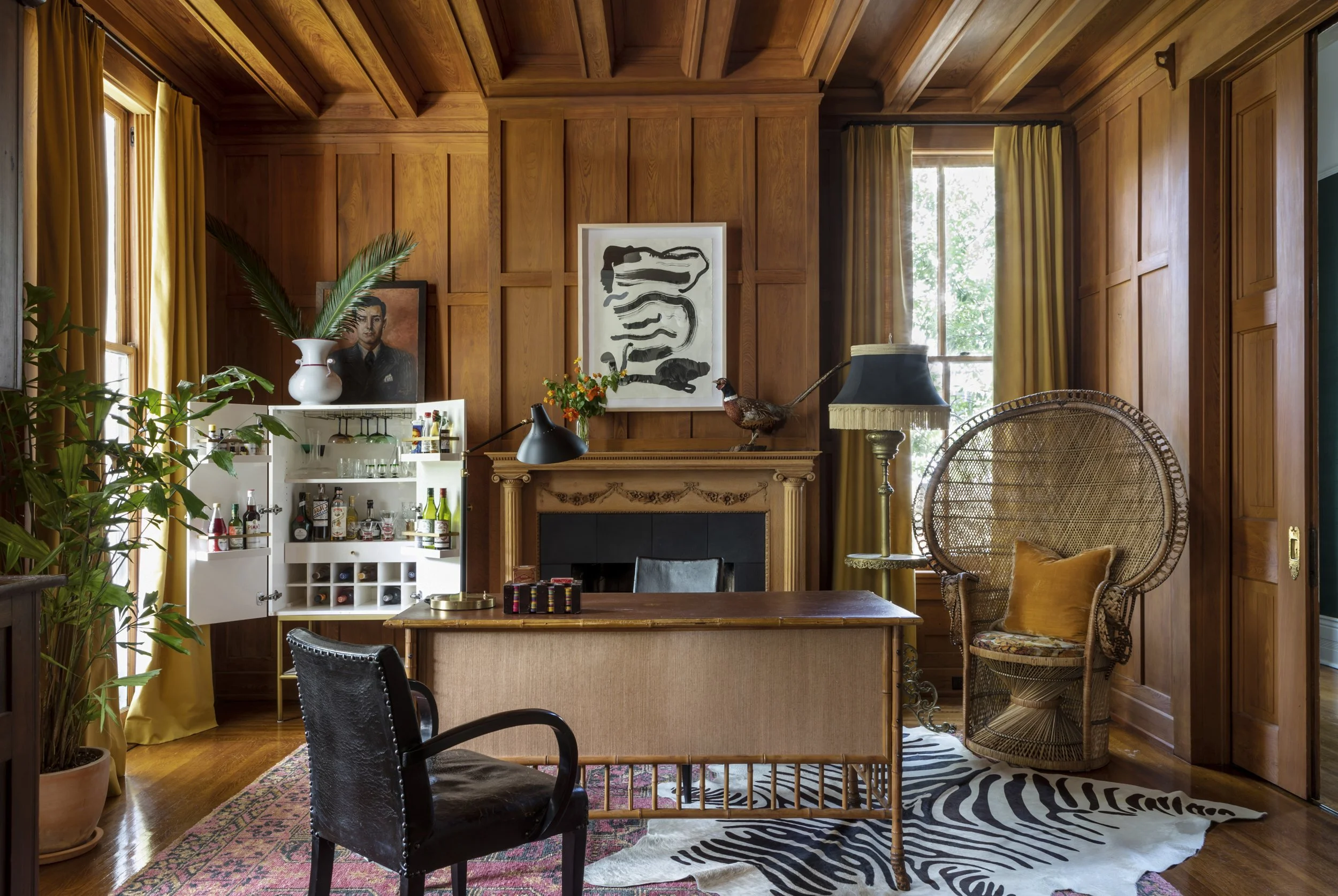 A cozy living room with wooden paneled walls and ceiling, decorated with artwork, plants, and vintage furniture, including a rattan chair and a small bar with bottles.