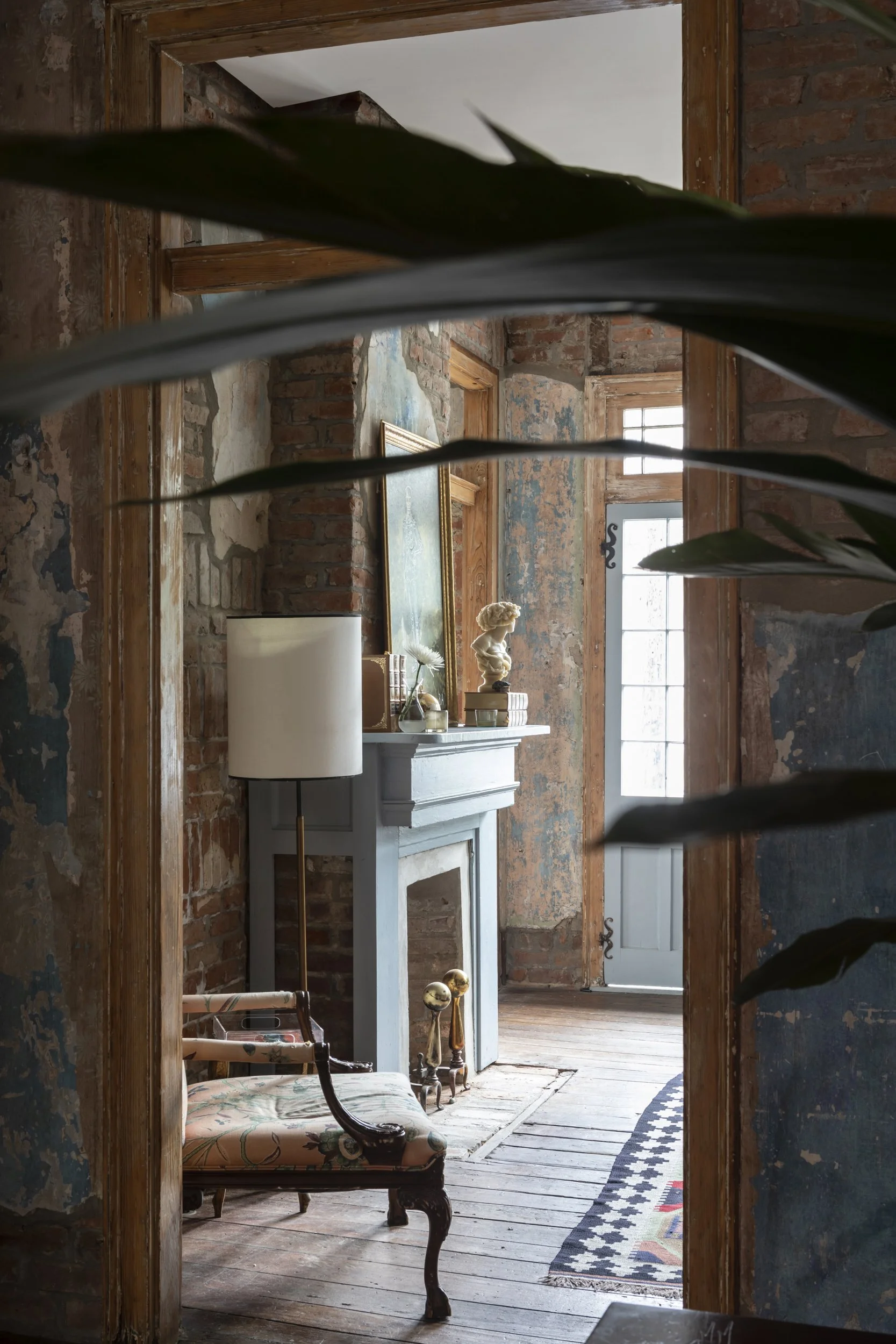 A view of a rustic interior room seen through a doorway with exposed brick walls, wooden trim, large windows, and vintage furniture including a floral upholstered bench and a white fireplace mantel decorated with art, a sculpture, and a lamp.