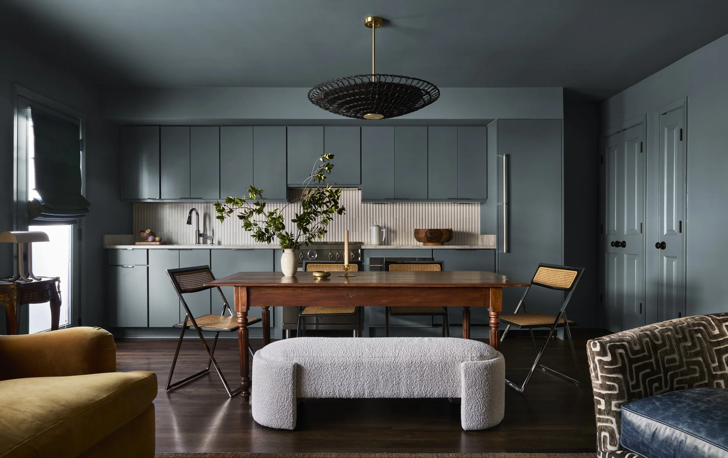 A modern kitchen and dining area with blue cabinets, a wooden dining table, and black metal chairs with woven seats. There is a potted plant on the table, a window with blinds, and a black pendant light fixture hanging from the ceiling.