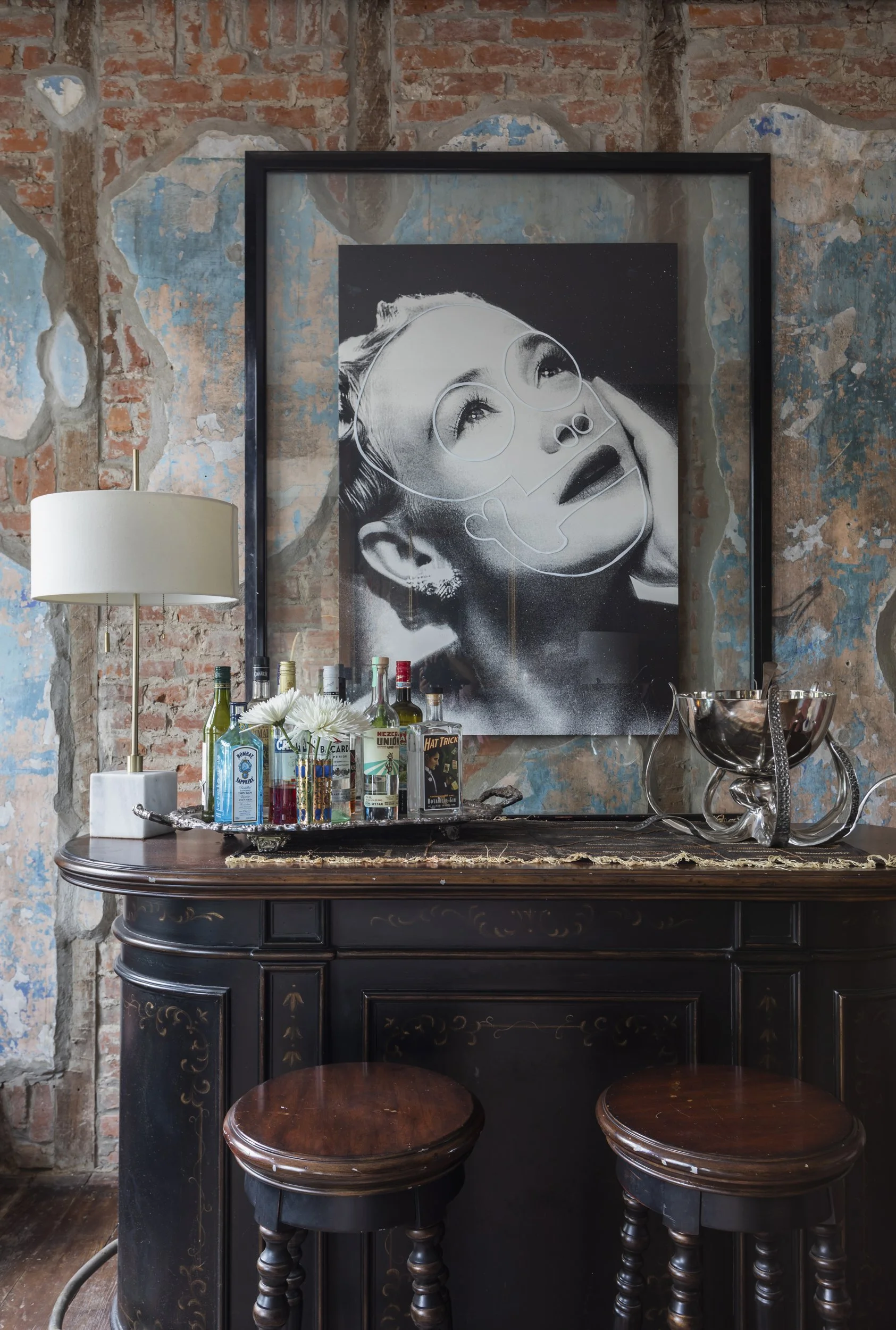 A bar or countertop against a rustic brick and painted wall, with various bottles of alcohol, a tray with flowers, a lamp, and a framed black and white portrait of a woman with glasses and makeup, with two wooden stools in front.