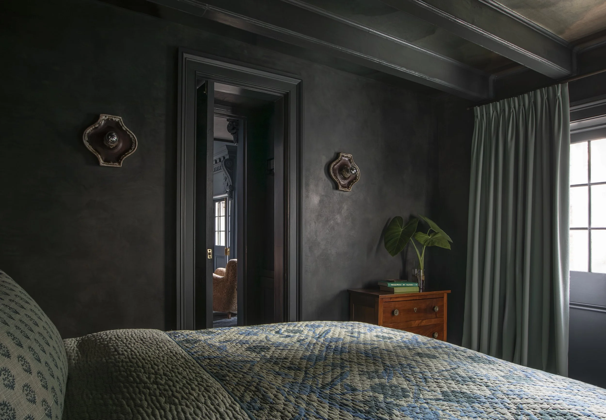A bedroom with dark green walls, a window with light-colored curtains, a wooden nightstand, and a bed with a patterned quilt. There are two vintage-style wall lamps and a green plant near the window.