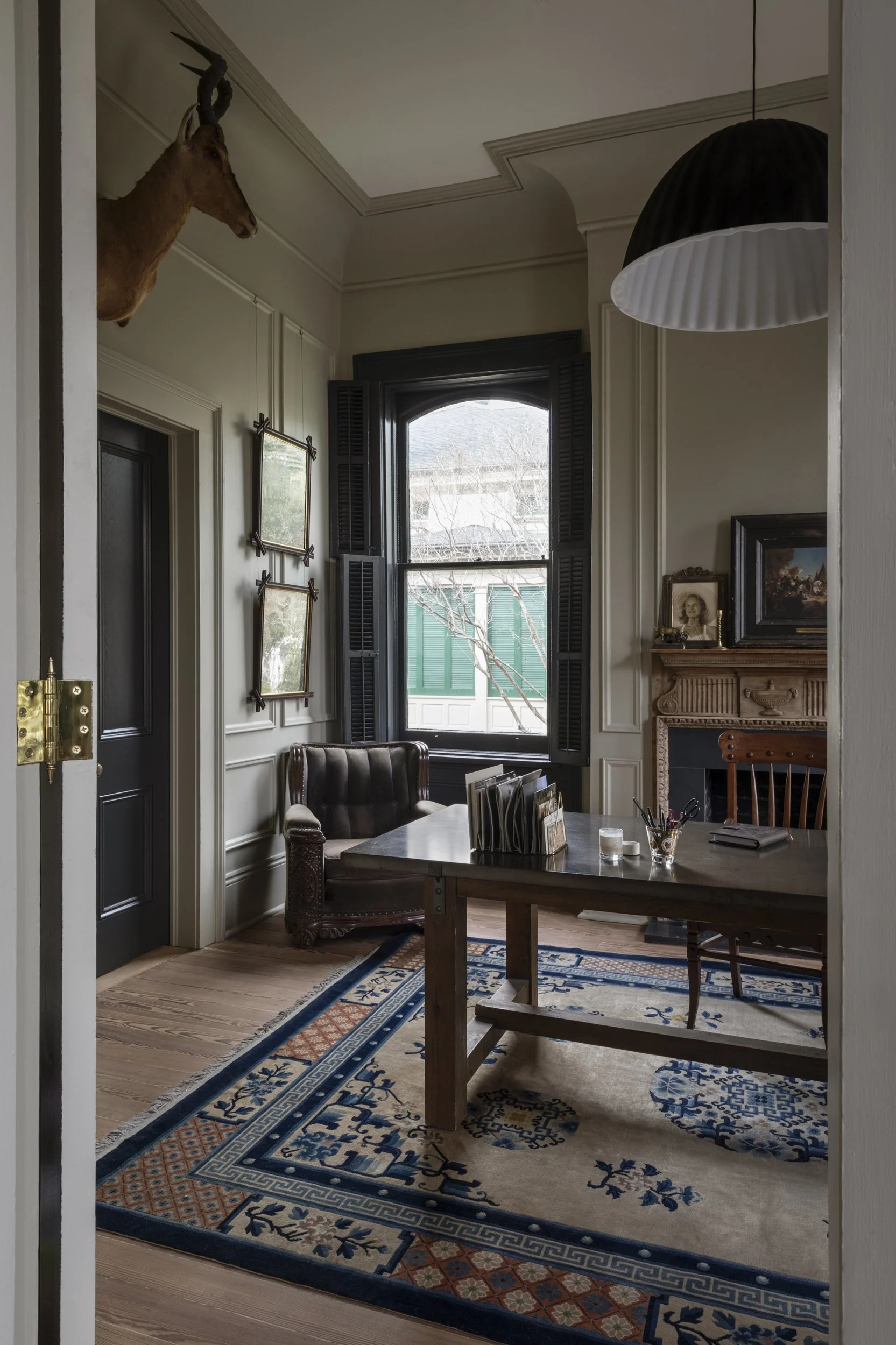 A cozy dining room with a wooden table, leather armchair, and a colorful patterned rug. There is a large window with black shutters, a fireplace with framed photos and artwork on top, and a mounted deer head on the wall. The room is decorated with fr