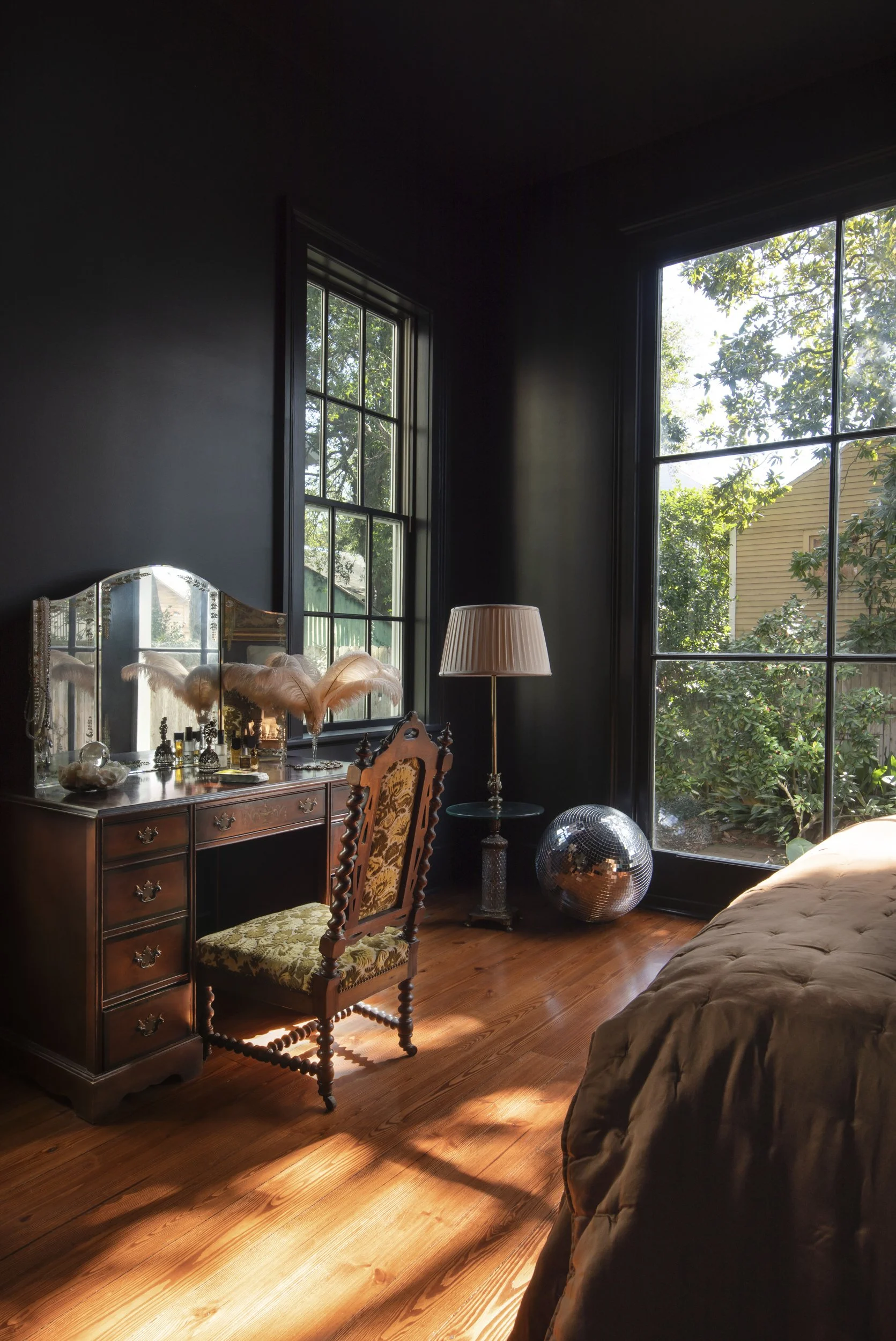 A cozy bedroom with hardwood floors, large windows letting in natural light, a vintage wooden dresser with a mirror, decorative feathers, perfume bottles, a lamp, an ornate chair with floral upholstered seat, and a bed with a beige coverlet.