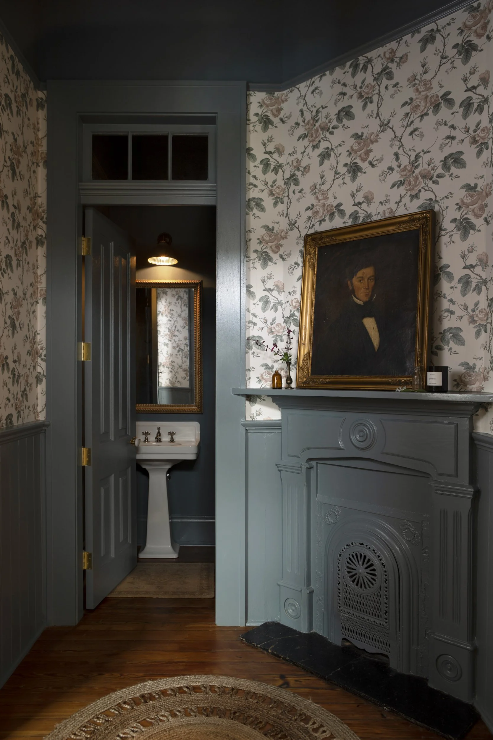 A cozy corner with a vintage portrait on a blue painted fireplace mantel, floral wallpaper, a mirror, and a small sink with gold fixtures in a homely setting.