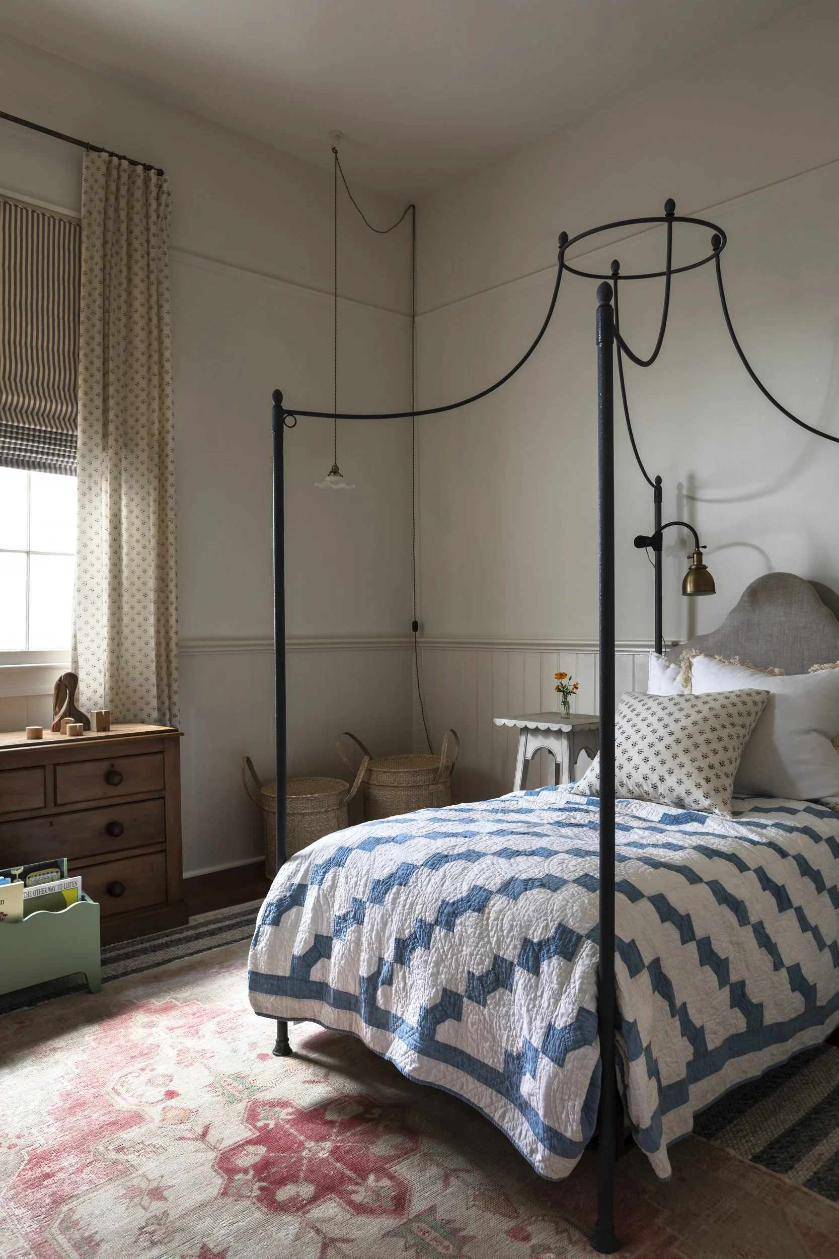A cozy bedroom featuring a black four-poster bed with a blue and white zigzag patterned quilt. There's a beige headboard with matching pillows, a side table with a small vase of orange flowers, a window with striped and floral curtains, a wooden dres