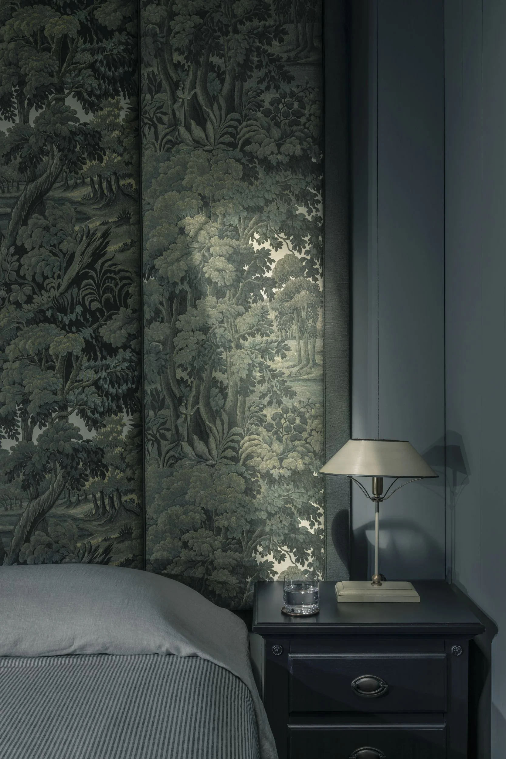 A bedroom with a bed, a blue-painted wall, a black nightstand with drawers, and a lamp and glass of water on it. There is a large painted mural of trees on the wall behind the bed.