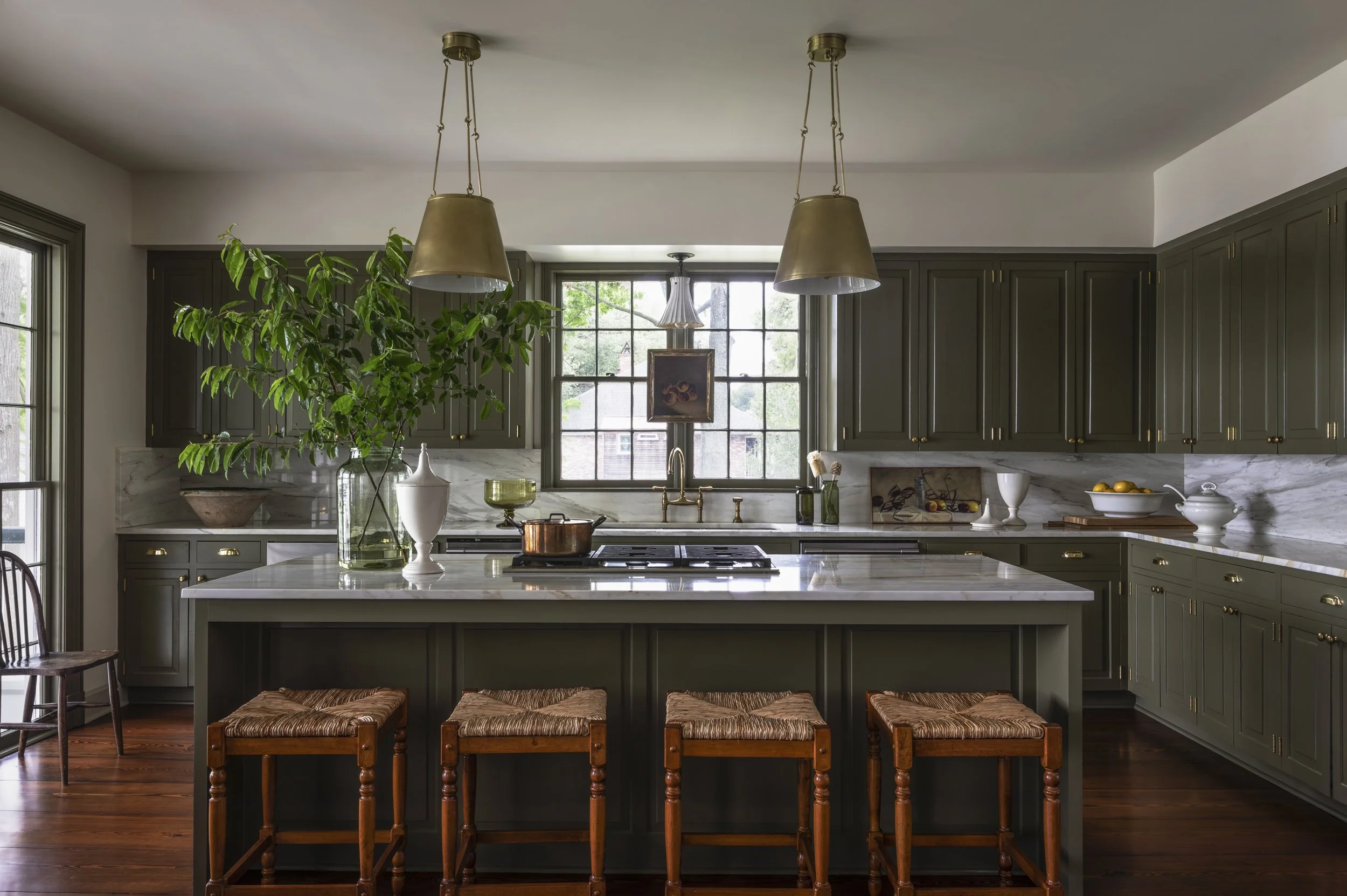 A modern kitchen with a white marble island, dark green cabinets with gold handles, and large windows. Hanging above the island are two gold pendant lights. On the countertop are decorative vases, a copper pot, a painting, and a bowl of lemons. Four 