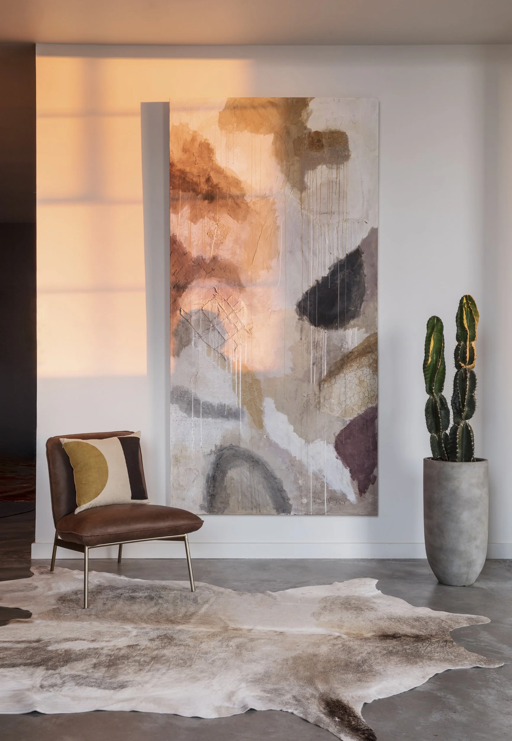 Modern interior with abstract wall art, a brown leather chair with a yellow and brown pillow, a large potted cactus, and a cowhide rug on a concrete floor.