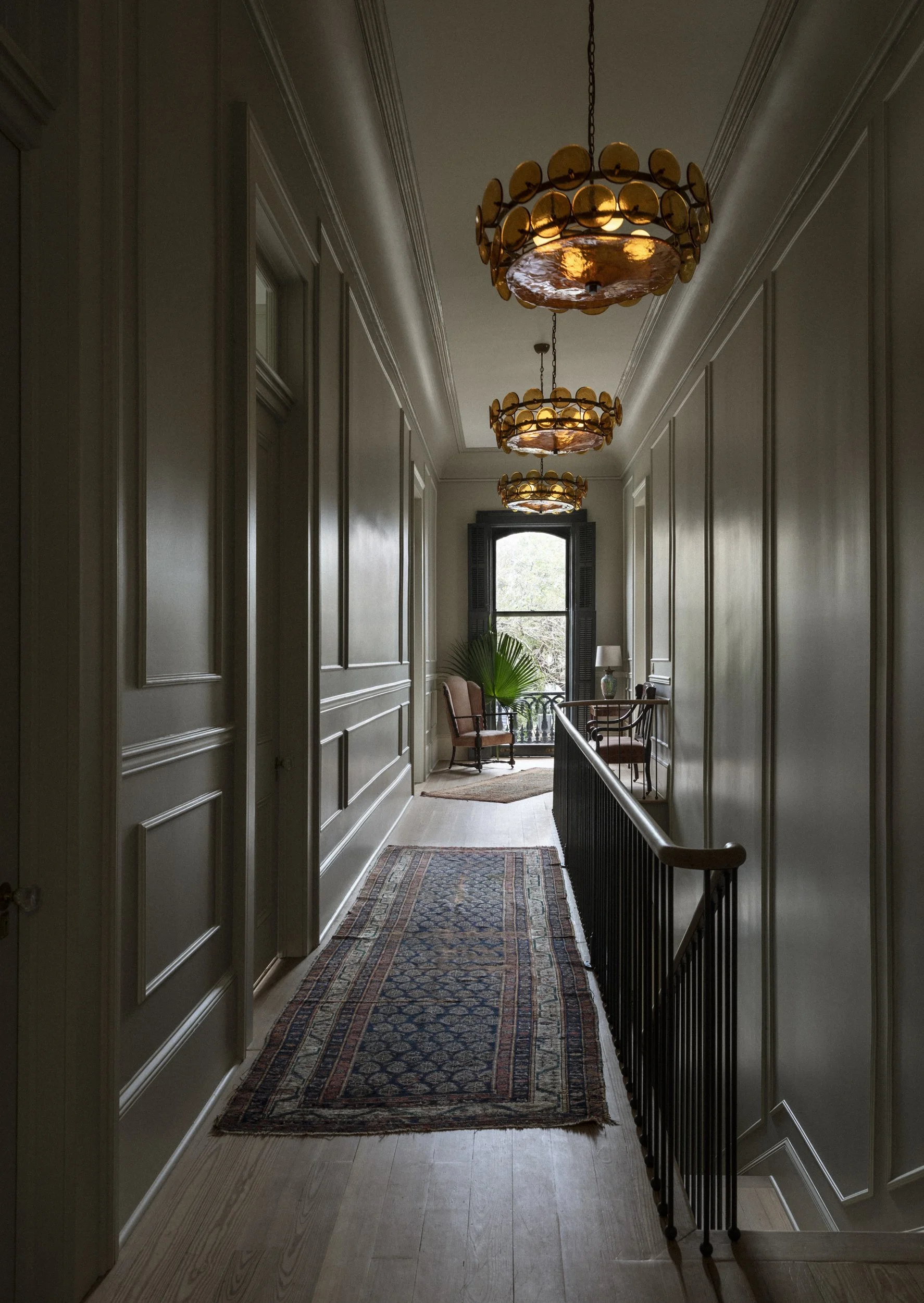 Long hallway in a house with white paneled walls, hanging circular chandeliers, a patterned runner rug, a window with black shutters at the end, and chairs by the window.