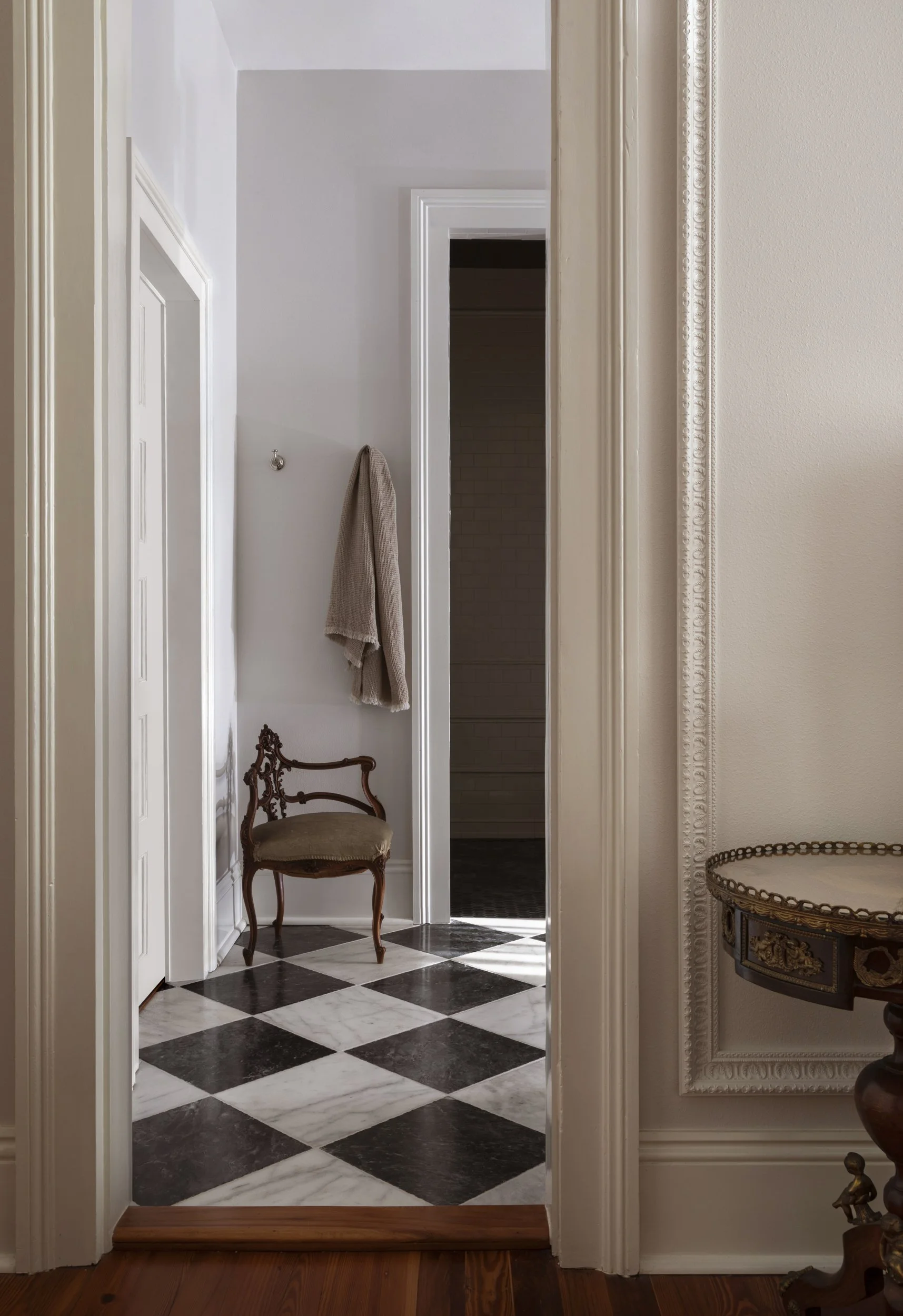 Interior view of a hallway with a black and white checkered marble floor, a vintage wooden chair with upholstered seat in front of a white wall with decorative molding, and a towel hanging on a hook on the wall. There is a doorway leading to a dark t