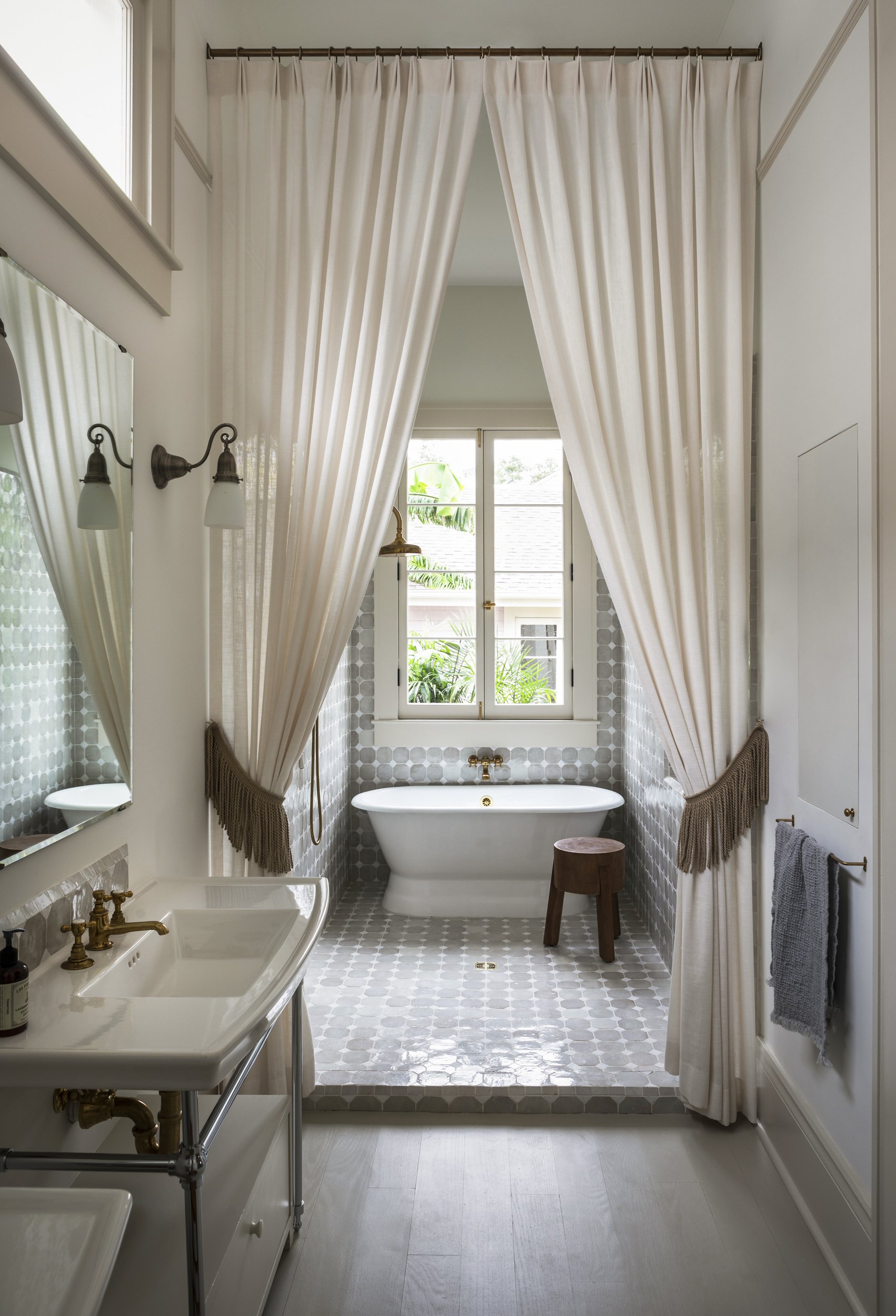 A vintage-style bathroom with a freestanding bathtub beneath a large window with cream curtains, a small round wooden stool, a white sink with brass fixtures, a mirror, wall-mounted light fixtures, and gray towels.