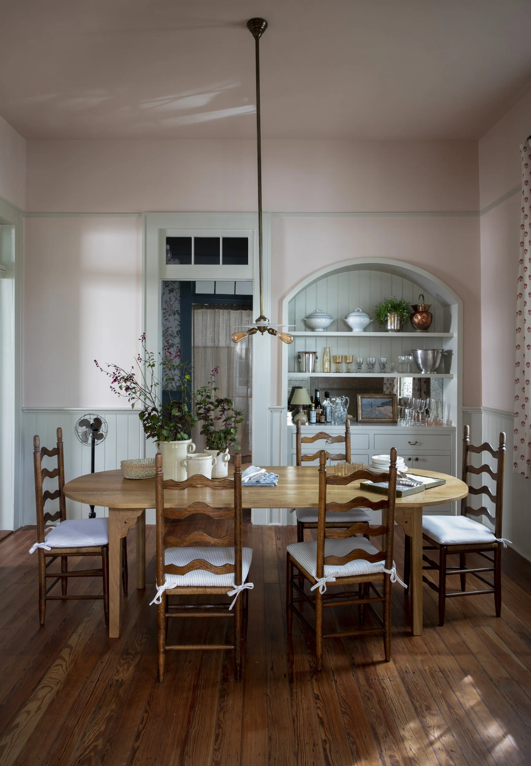 A dining room with a wooden table surrounded by six wooden chairs with white cushion seats. On the table are various white and cream-colored vases, a small woven basket, and some napkins. In the background, a built-in cream-colored cabinet with glass
