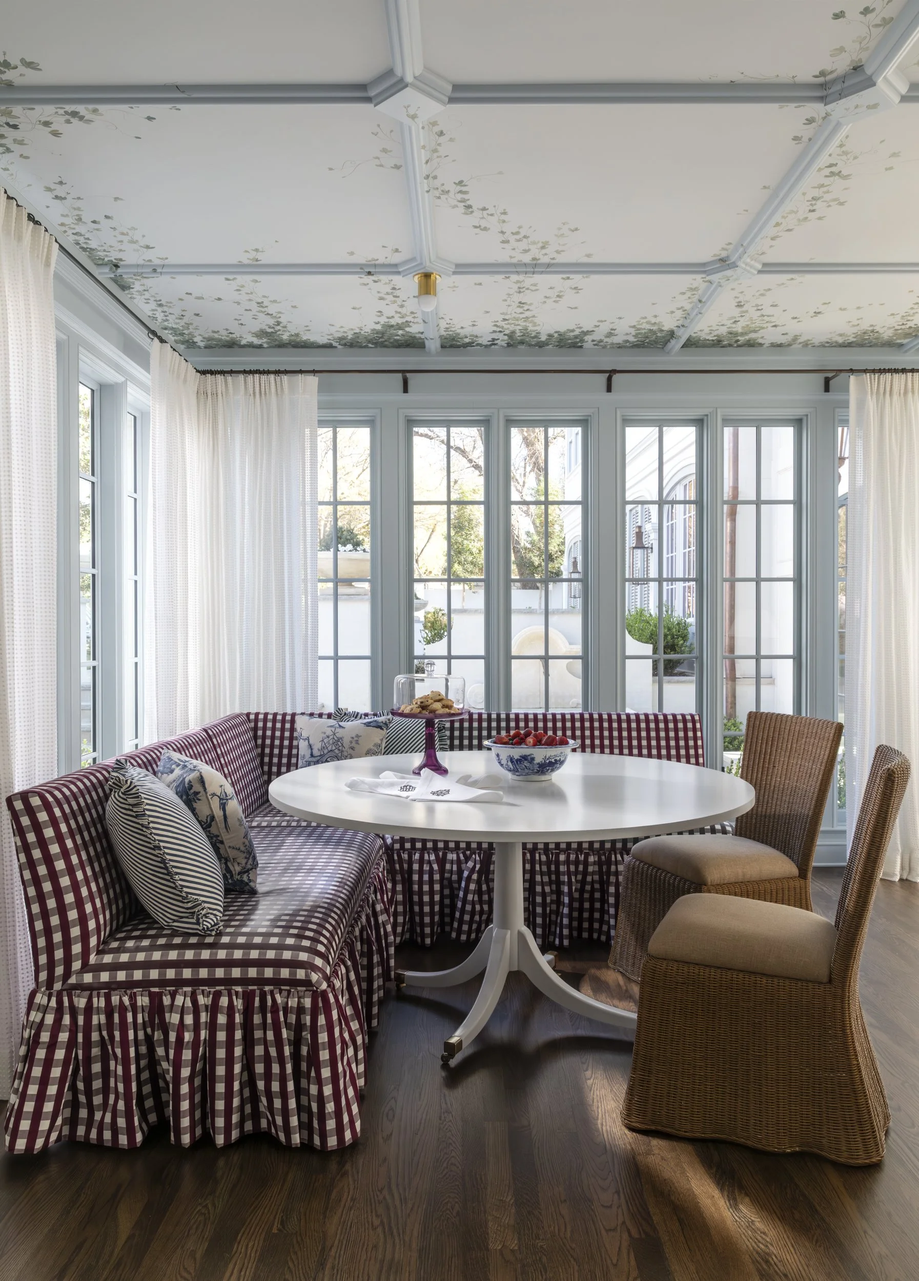 A dining room with a white round table, surrounded by a red checkered bench with pillows and wicker chairs, large windows with cream curtains, revealing outdoor greenery, ceiling with floral design, dark wood flooring.