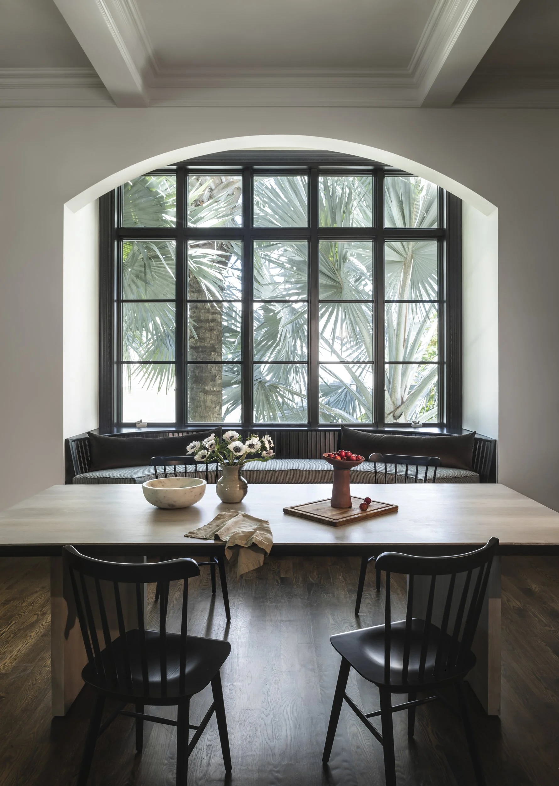 Dining room with a large window showing palm trees outside, a light wooden table with a vase of white flowers, a bowl, a cloth, and a centerpiece with cherries, surrounded by black chairs, with a bench along the window.