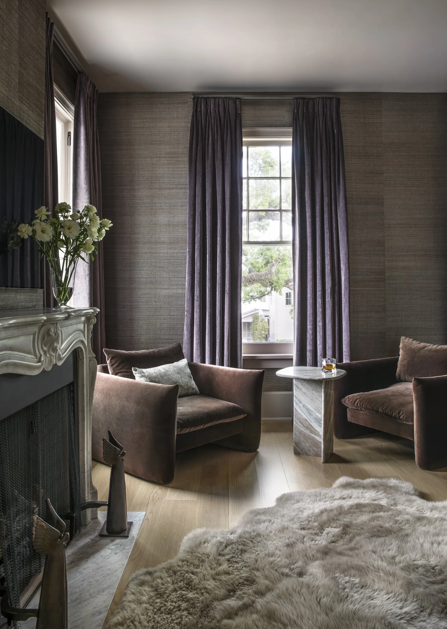 A cozy living room with a large window covered by purple curtains, a plush brown velvet sofa with pillows, a marble side table with a glass of drink, a decorative fireplace with flowers, a faux animal fur rug, and wooden flooring.