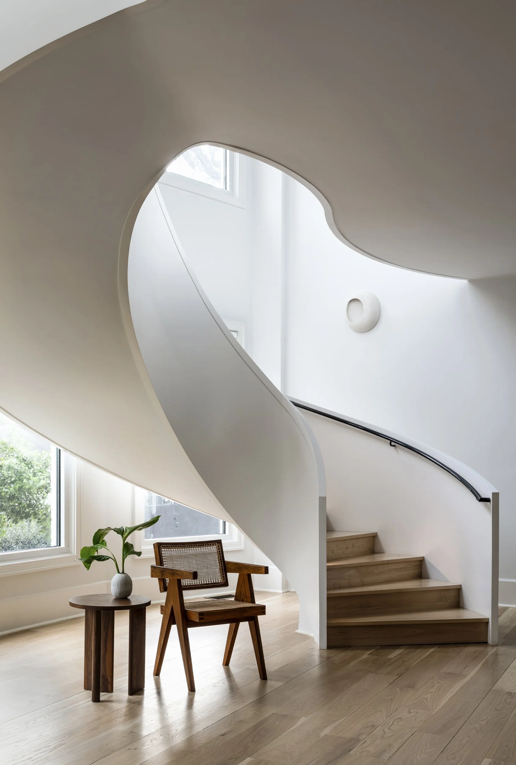 Modern interior with a curved staircase, large windows, a small wooden table with a green potted plant, and a wooden chair in a bright room.