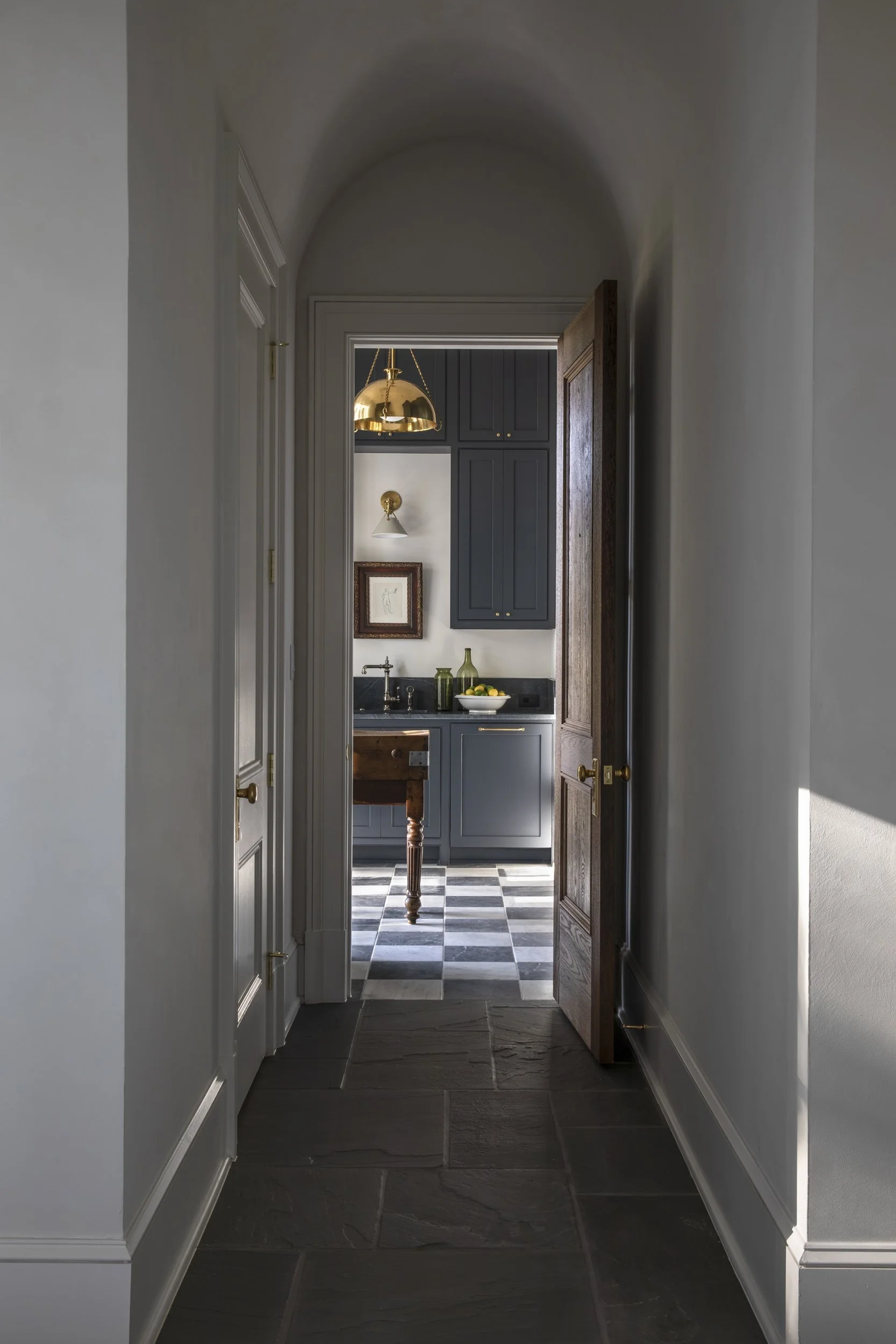 View through a narrow hallway into a small kitchen area with dark blue cabinets, a checkered black and white floor, and gold-colored lighting fixtures.