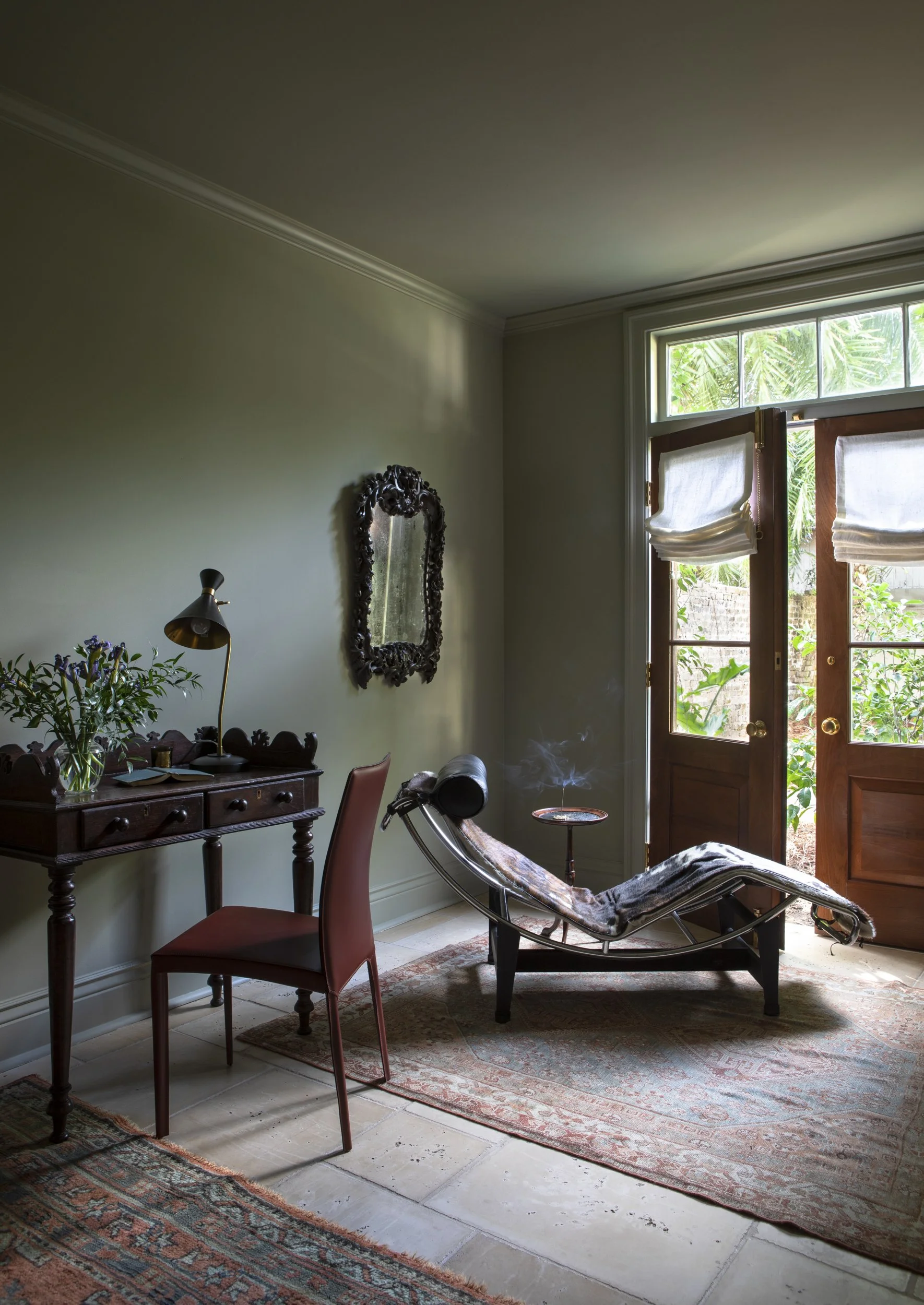 Interior view of a room with a wooden lounge chair with a fur blanket, a small side table with a lit incense holder, a vintage mirror on the wall, a desk with a lamp and flowers, a red chair, and an open wooden door with blinds, revealing a garden ou