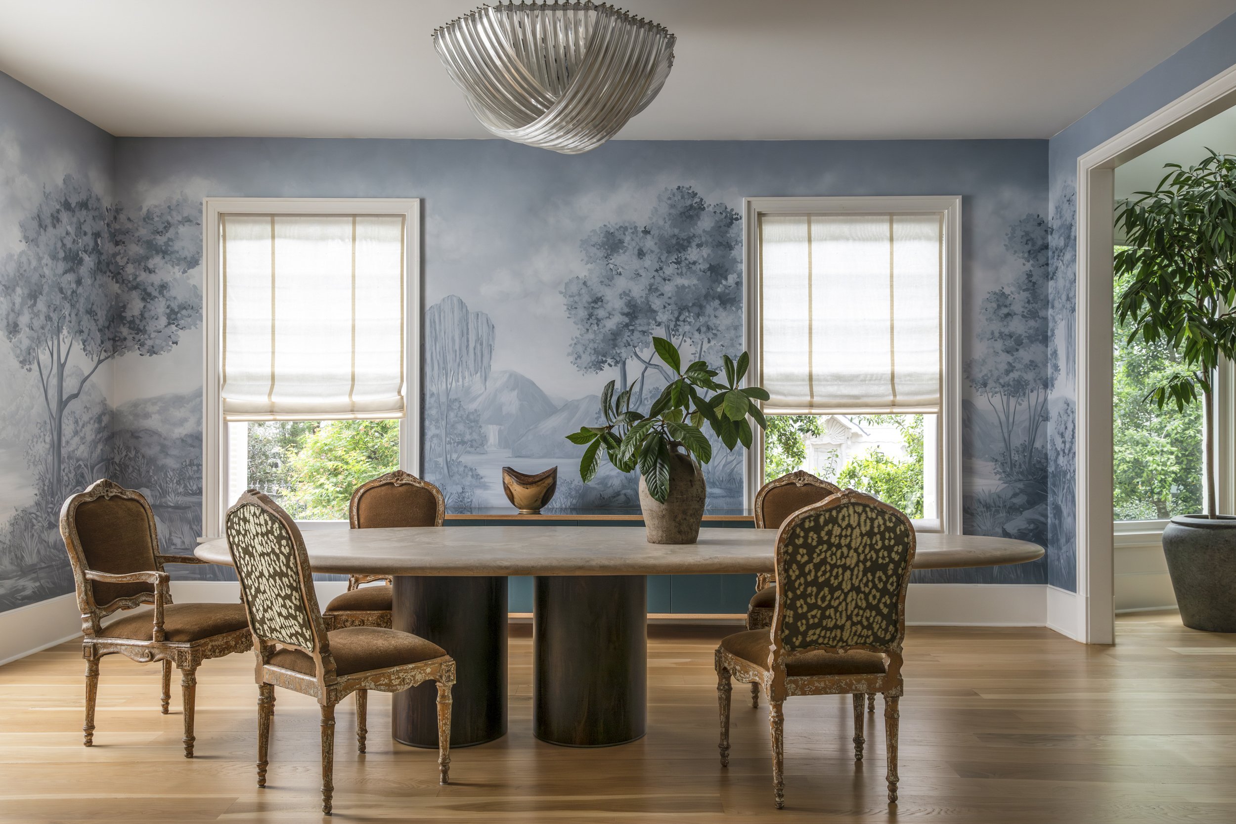A dining room with a large round table surrounded by antique-style chairs with patterned upholstery. There are three large windows with white blinds, and a painted mural of trees and mountains on the wall behind the table. A large potted plant is on 