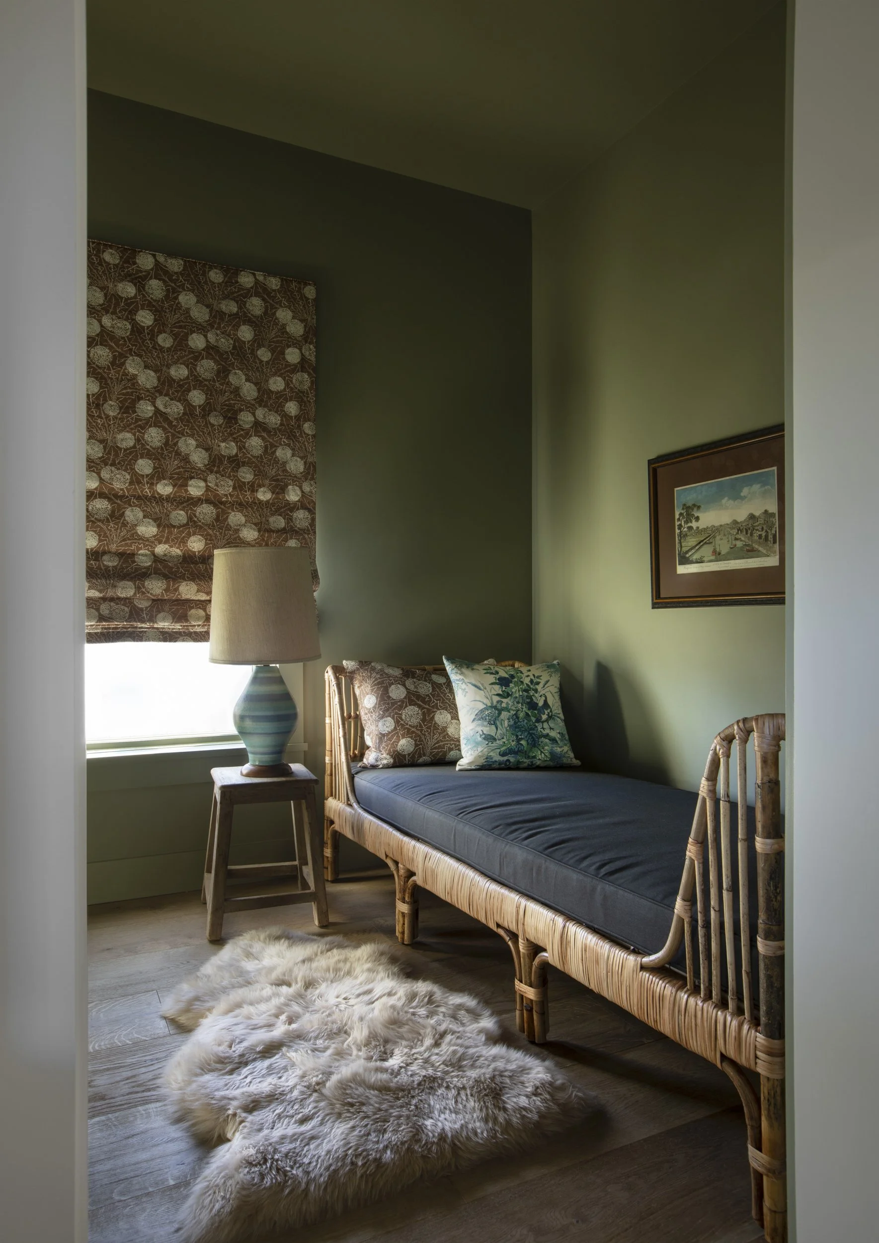 A cozy bedroom with a bamboo daybed, dark gray mattress, and decorative pillows featuring green and floral patterns. There's a wooden side table with a striped ceramic lamp, a window with a patterned brown and cream curtain, framed artwork on the gre