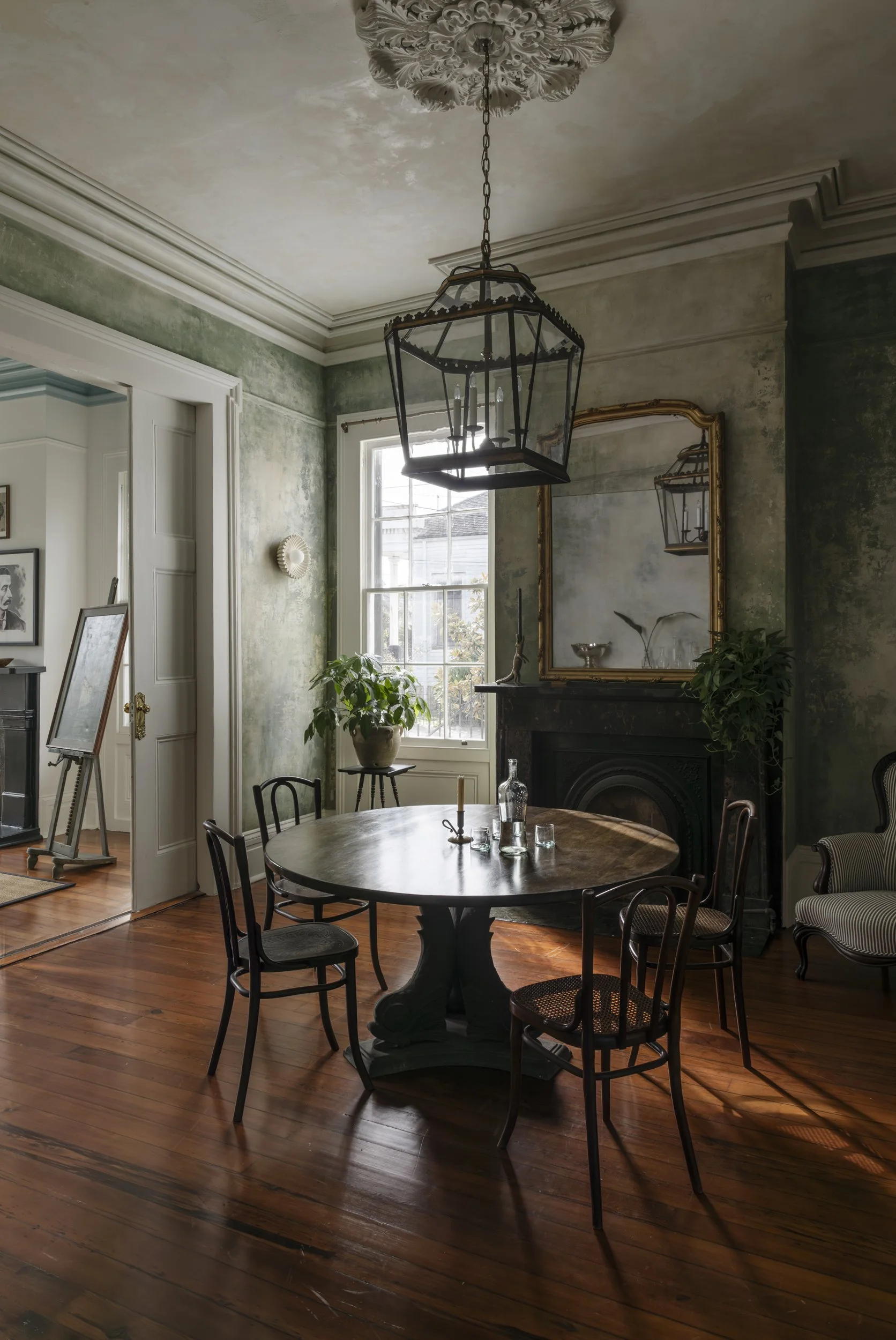 A cozy dining room with a round wooden table surrounded by six black chairs, a black fireplace with a gold-framed mirror above it, a window letting in natural light, green walls with a weathered finish, and a large outdoor view. A chandelier hangs fr