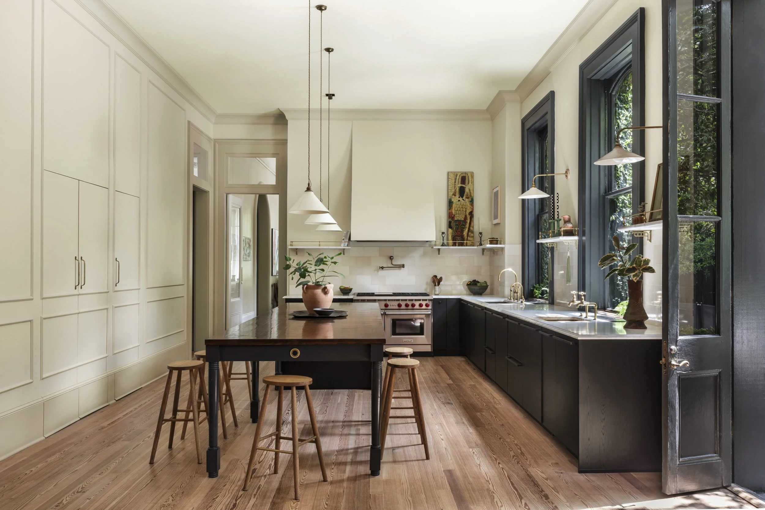 A bright kitchen with large windows, a wooden island with stools, white cabinets, black lower cabinets, a stove, and decorative items, illuminated by hanging pendant lights.