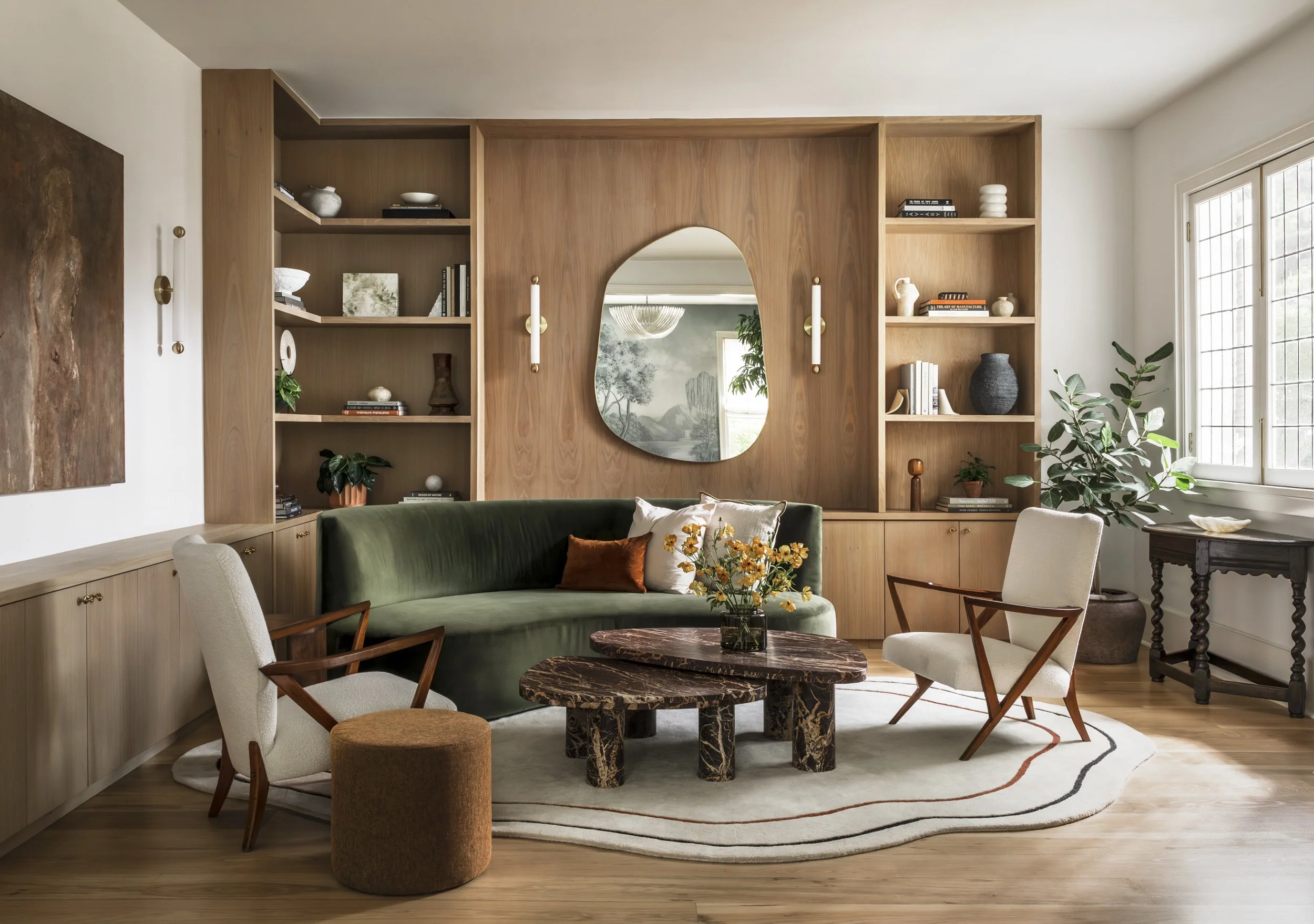 A living room with a green velvet sofa, beige chairs, a marble coffee table, built-in wooden shelves with decorative objects, books, potted plants, a round mirror, and large windows letting in natural light.