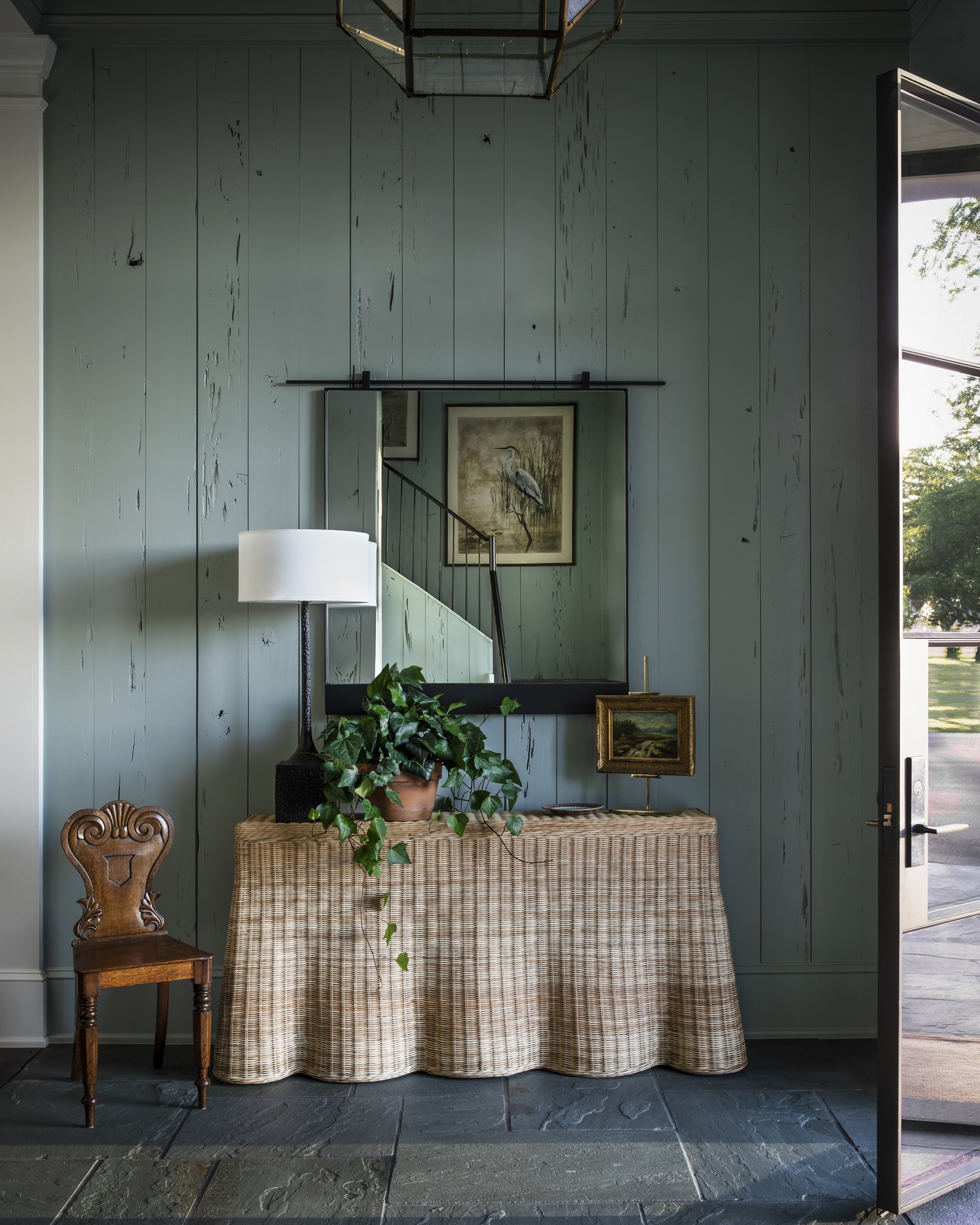Interior of a house with a green vertical wooden panel wall, a wicker table with a potted ivy, a vintage wooden chair, and framed artwork of a heron, with an open door and a large window showing trees outside.