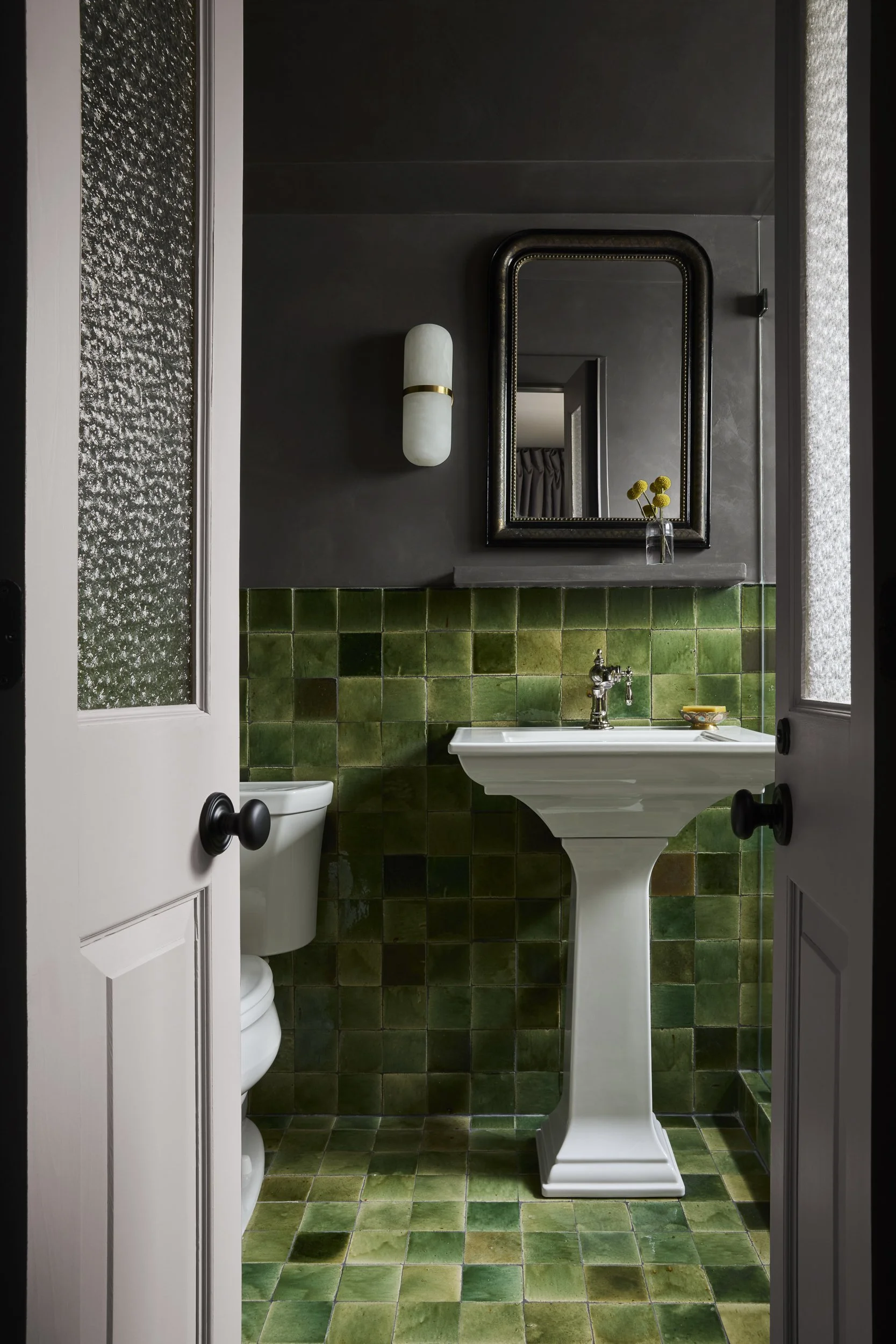 A bathroom with green tiled walls and floor, a white pedestal sink, a large mirror with a dark frame, a white wall sconce, a small vase with yellow flowers, and a toilet partially visible behind the door.