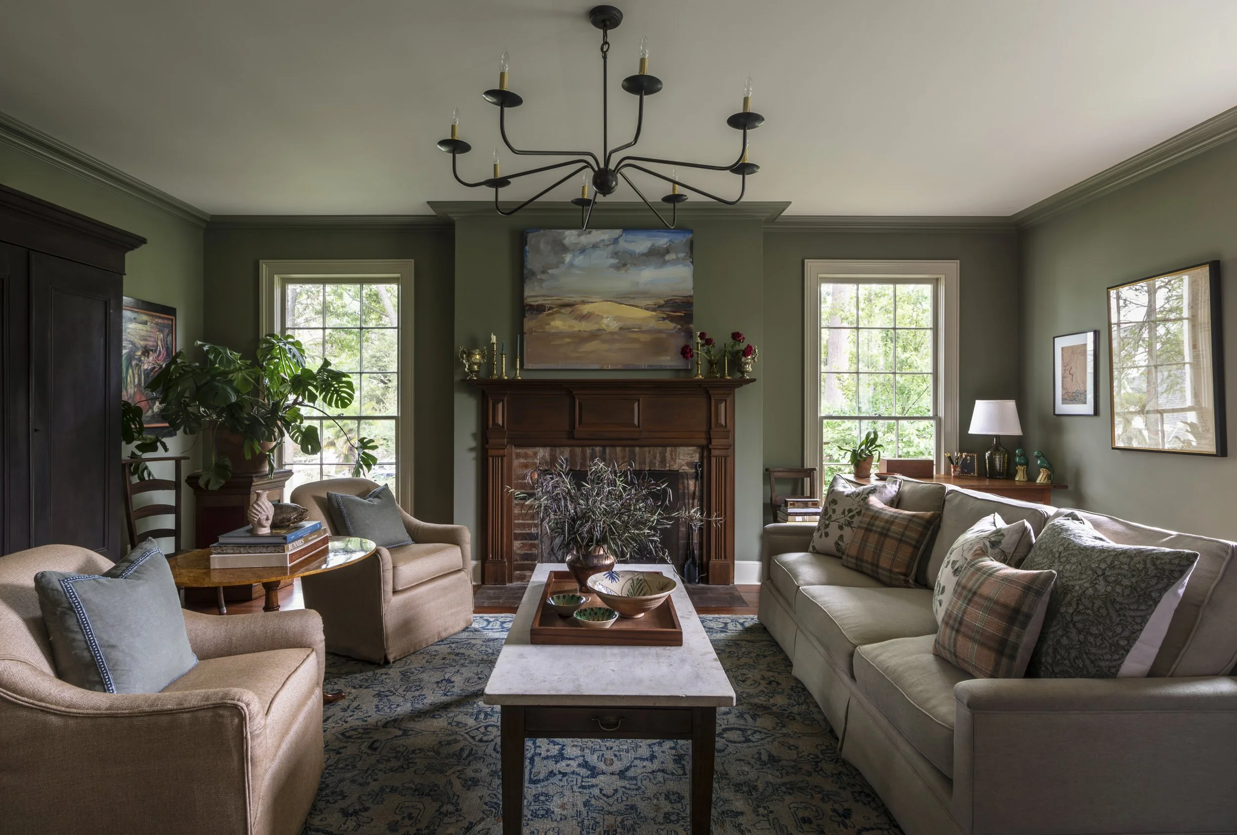 A cozy living room with green walls, large windows, a brick fireplace with a wooden mantel, a black chandelier, and seating including a beige armchair and a green sofa with plaid and floral cushions, decorated with plants, artwork, and a coffee table