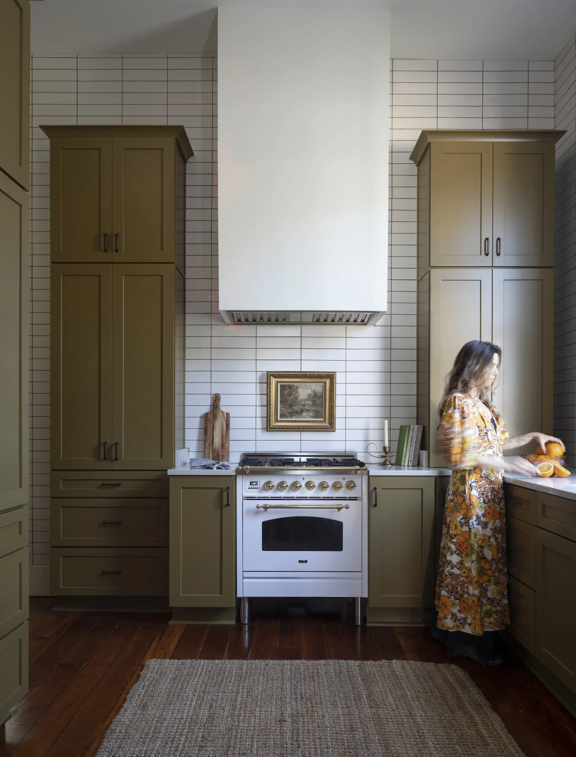 A woman in a floral dress preparing food in a kitchen with green cabinets, a white stove with brass handles, a framed landscape painting, and a beige rug on dark wooden floors.