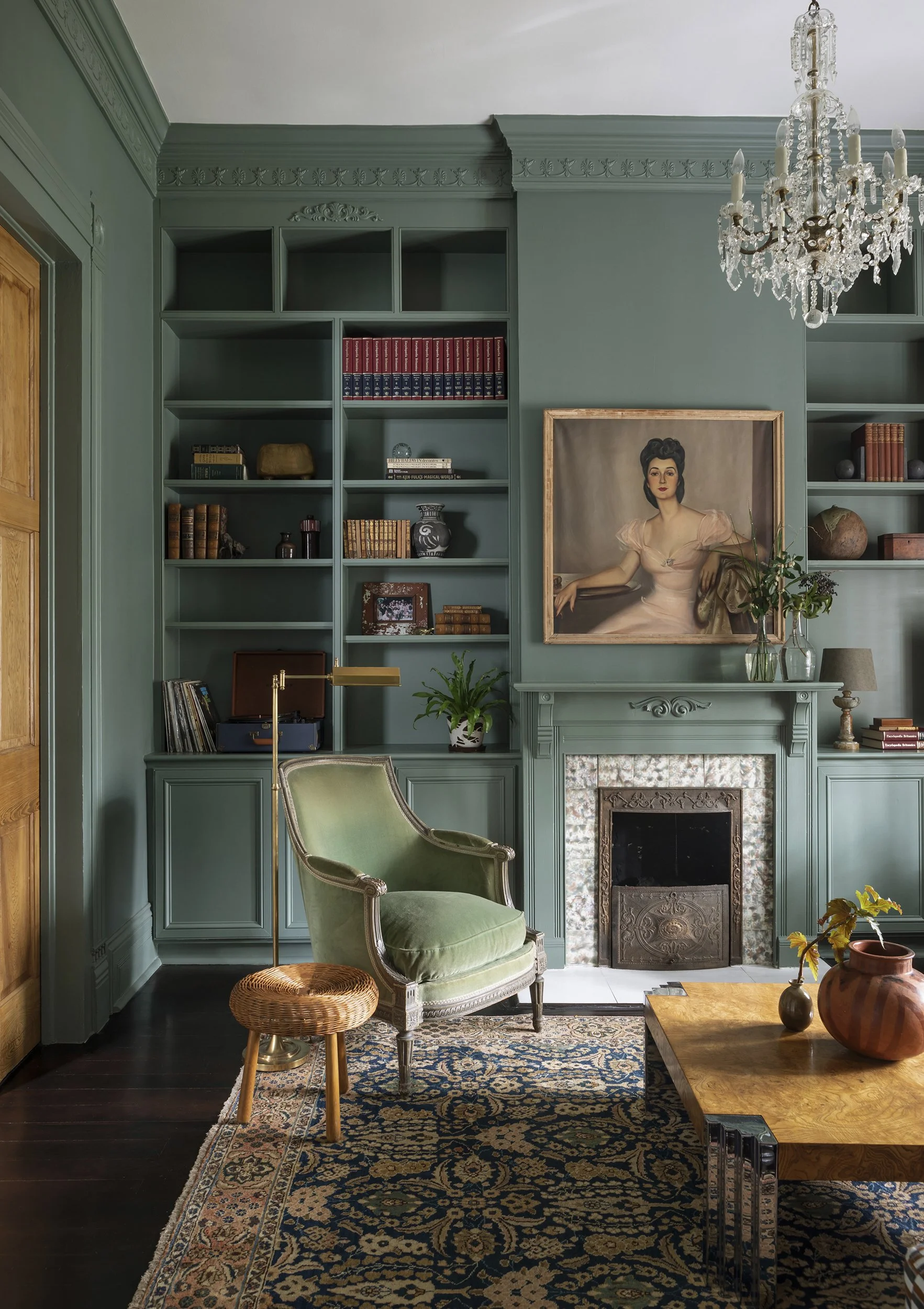 A vintage living room with green walls, a fireplace, a large oil painting of a woman, a green armchair, a small wicker stool, a wooden coffee table with vases, a patterned rug, shelves filled with books and decorative objects, and a crystal chandelie